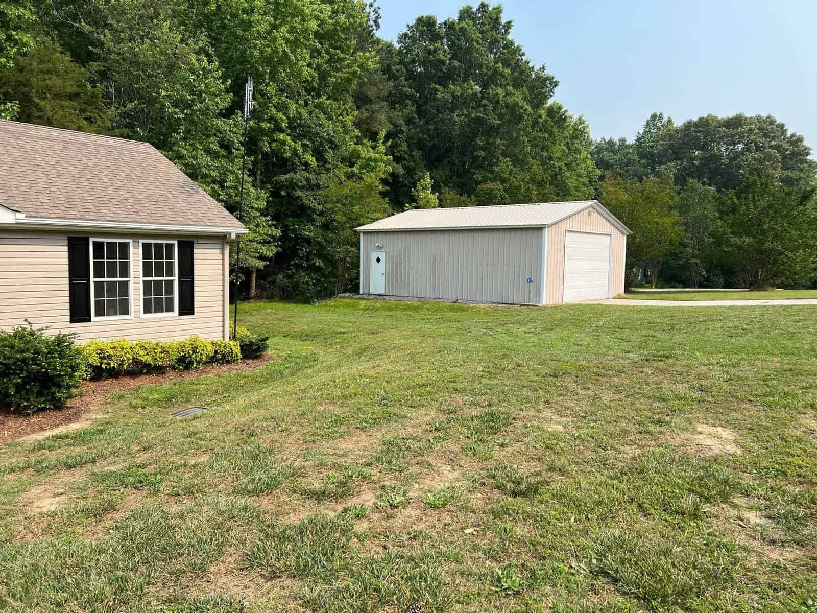 A small, tan house with black shutters and a brown roof is surrounded by green grass and bushes. To the right, a gray metal shed with a white door and garage entrance sits against a backdrop of trees under a blue sky.