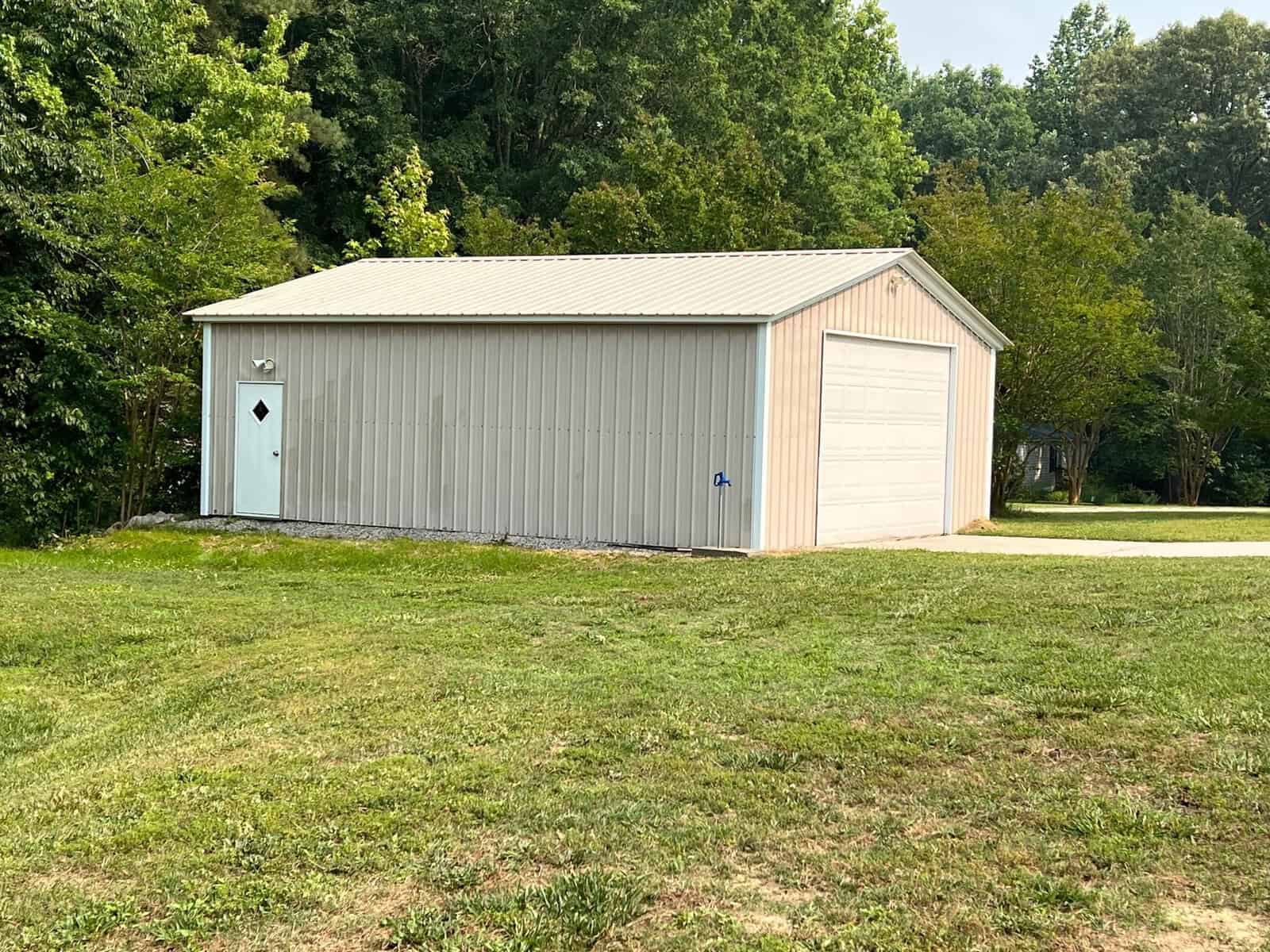 A beige metal storage building with a white door and a large garage door sits on a grassy plot. Lush green trees surround the area, and a paved path leads to the garage entrance.