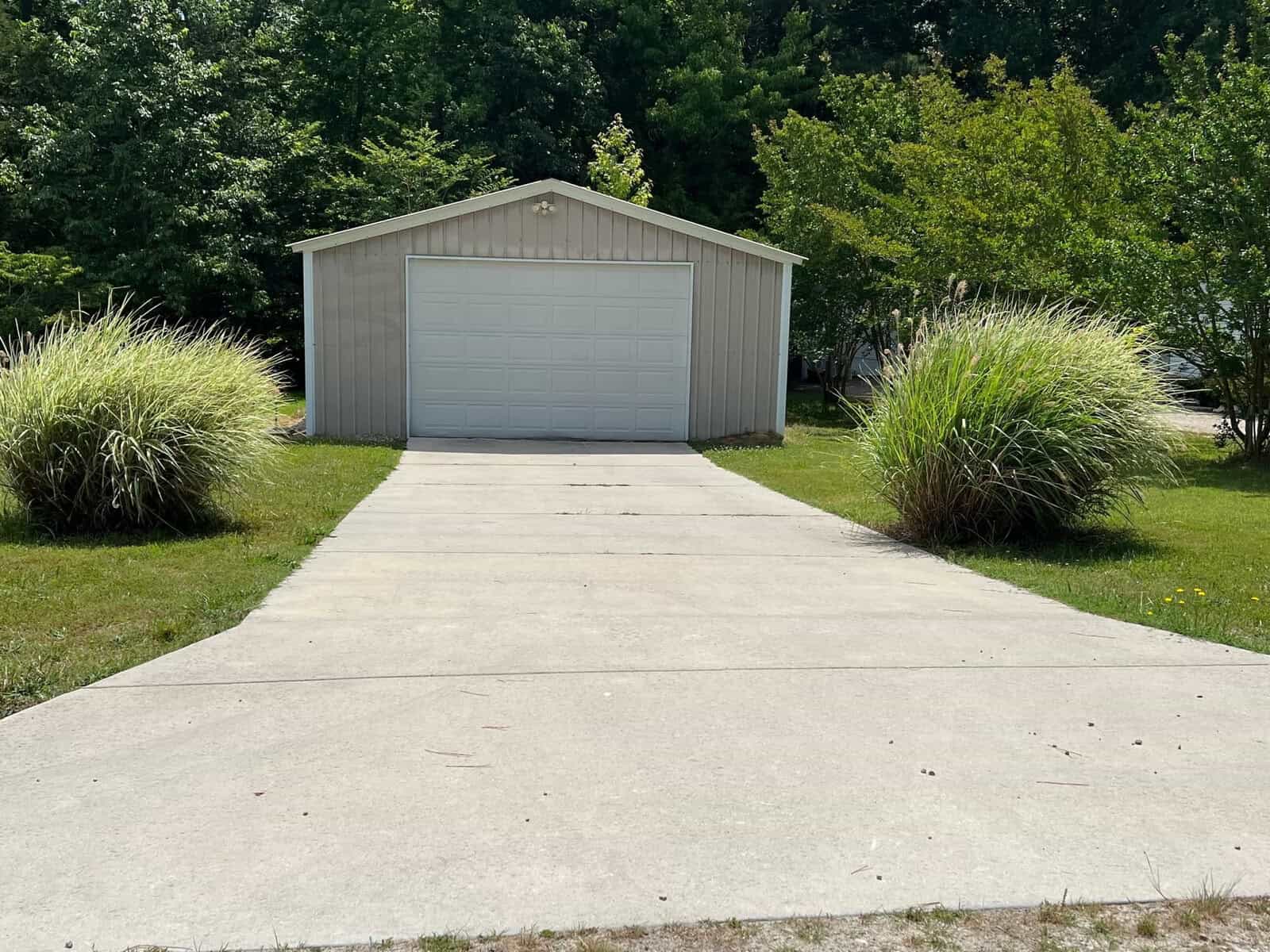 A small beige shed with a white garage door is at the end of a concrete driveway. Two large, green ornamental grasses are on either side of the driveway. The background features trees with lush green leaves.