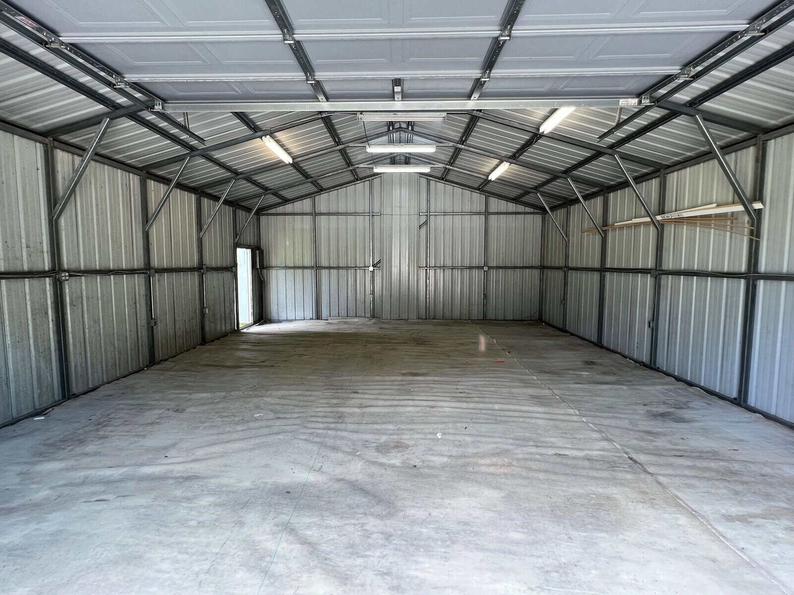 Interior of an empty metal garage with a concrete floor and open door at the back. The ceiling and walls are lined with metal panels, and fluorescent lights are installed overhead. The space is well-lit by natural light from the open door.