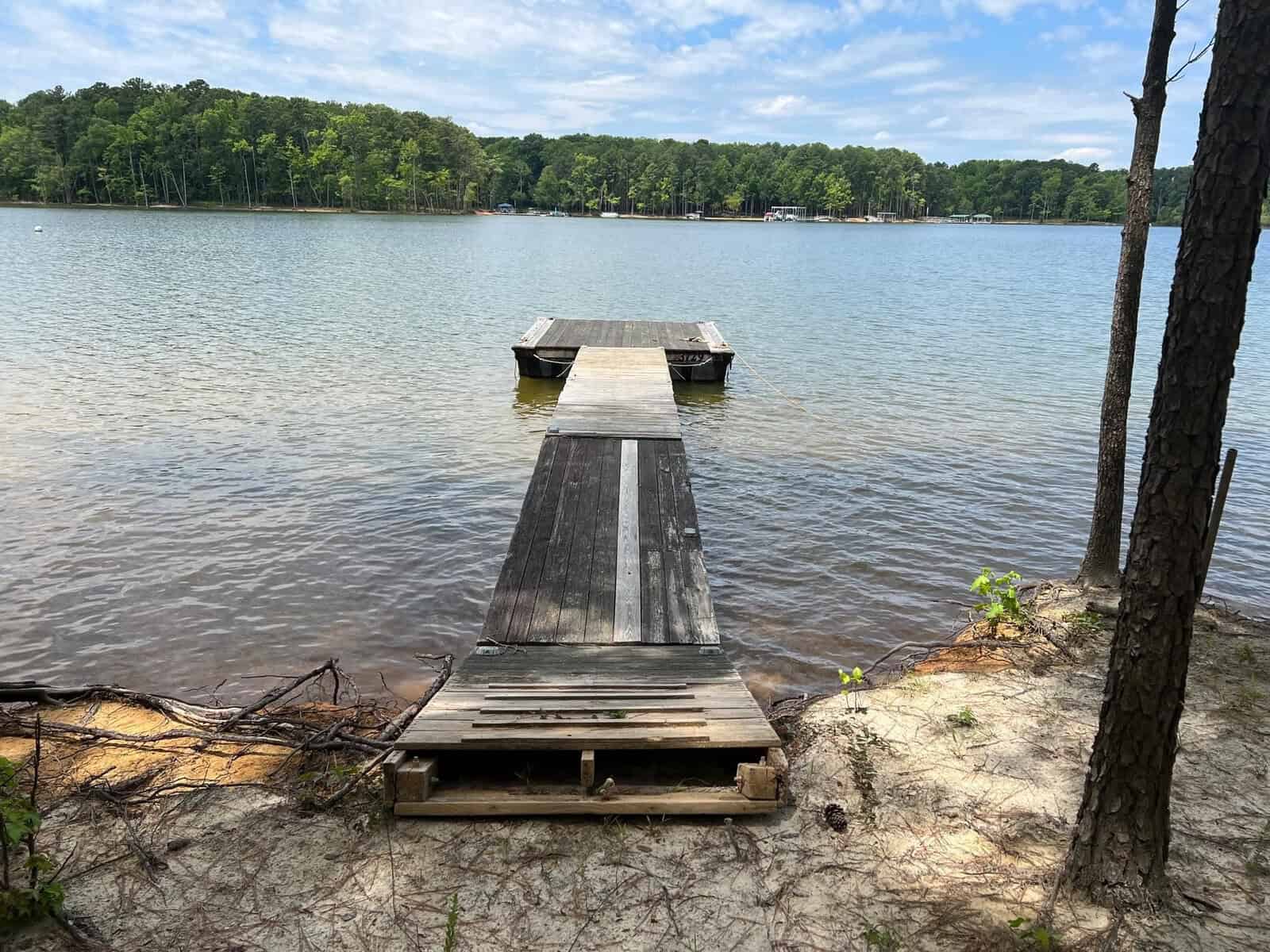 A wooden dock extends into a calm lake surrounded by lush green trees. The sky is partly cloudy, and the shoreline is sandy with patches of grass.
