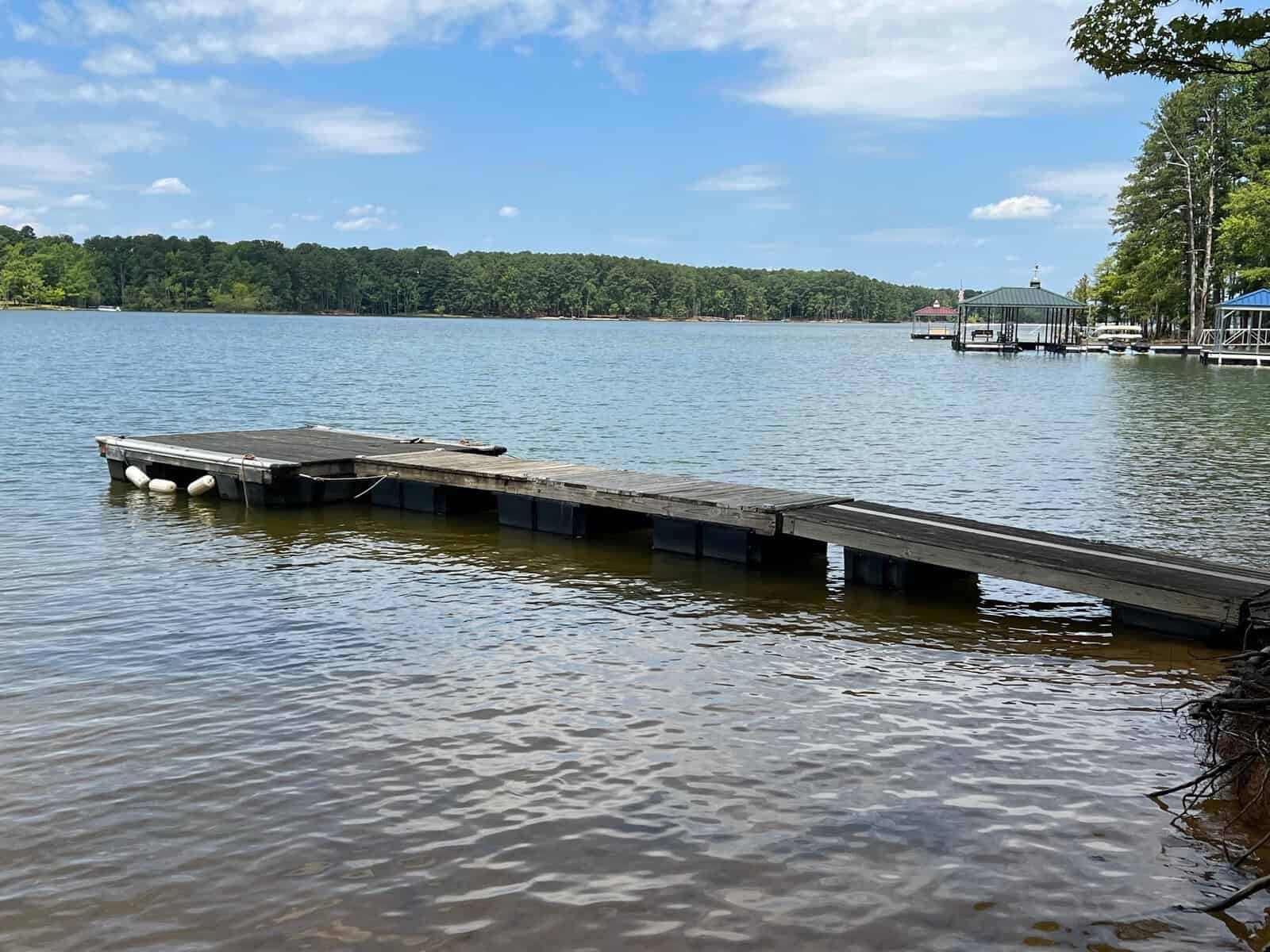 A calm lake with a wooden dock extending into the water. Trees line the distant shore under a partly cloudy sky, and there are small boat docks visible in the background.