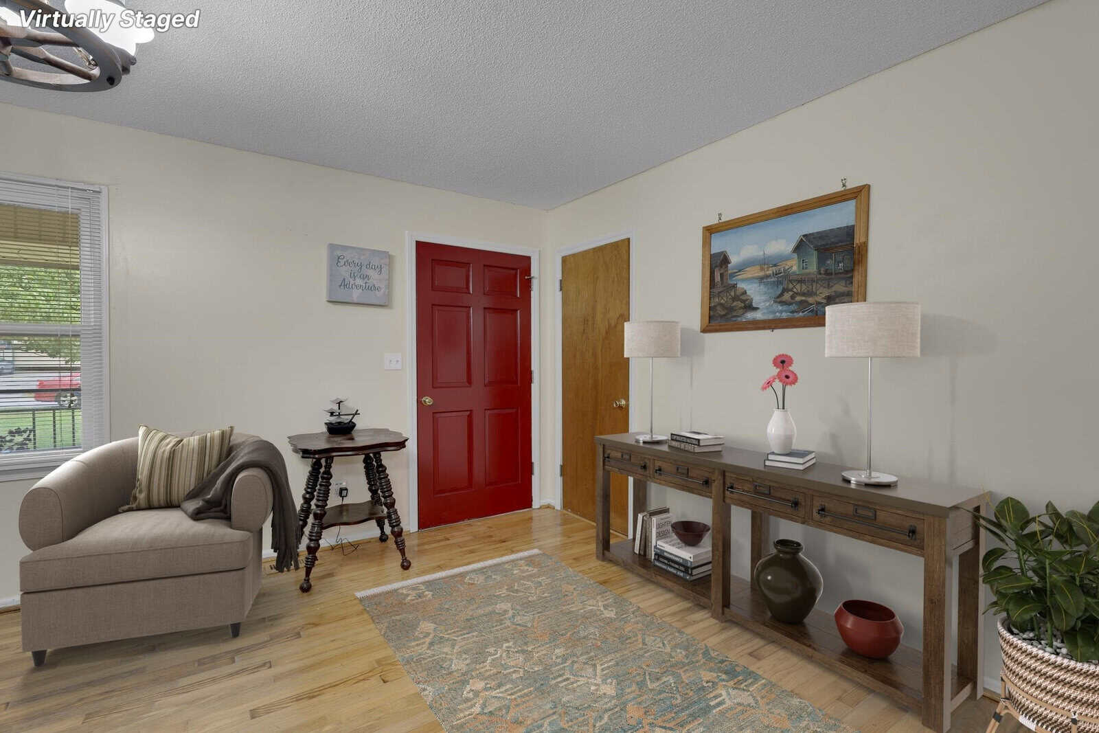 A neatly furnished living room featuring a tan sofa, a vintage-style round table, a wooden console table with two lamps, and a decorative rug, with a vibrant red door in the background. the room has a "family" themed wall decor and a painting of a ship, with a text overlay indicating the space is virtually staged.