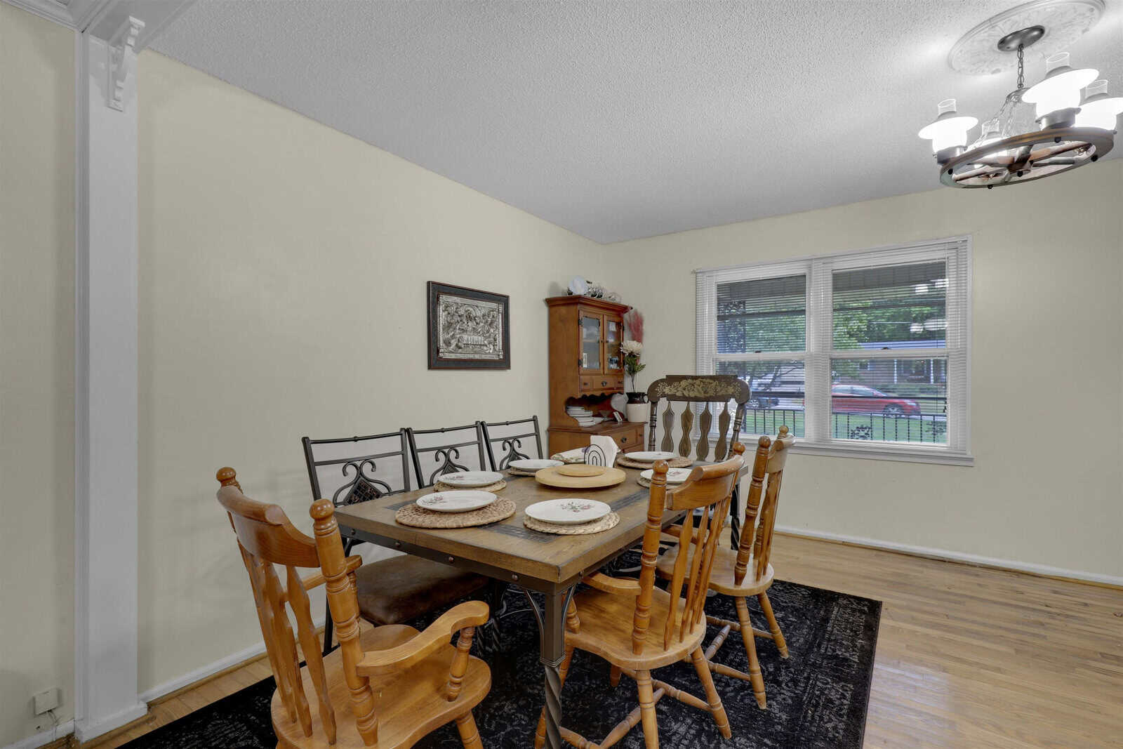 A well-lit dining room featuring a wooden table set for four, with matching chairs, a corner cabinet, and a picture on the wall; a window provides a view of the outdoors.