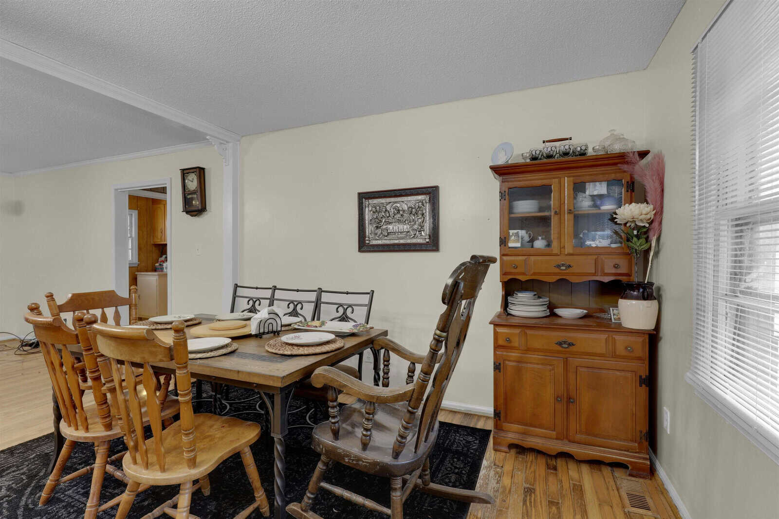A quaint dining room with a wooden table set for four, traditional chairs, and a vintage china cabinet next to a window with white blinds.