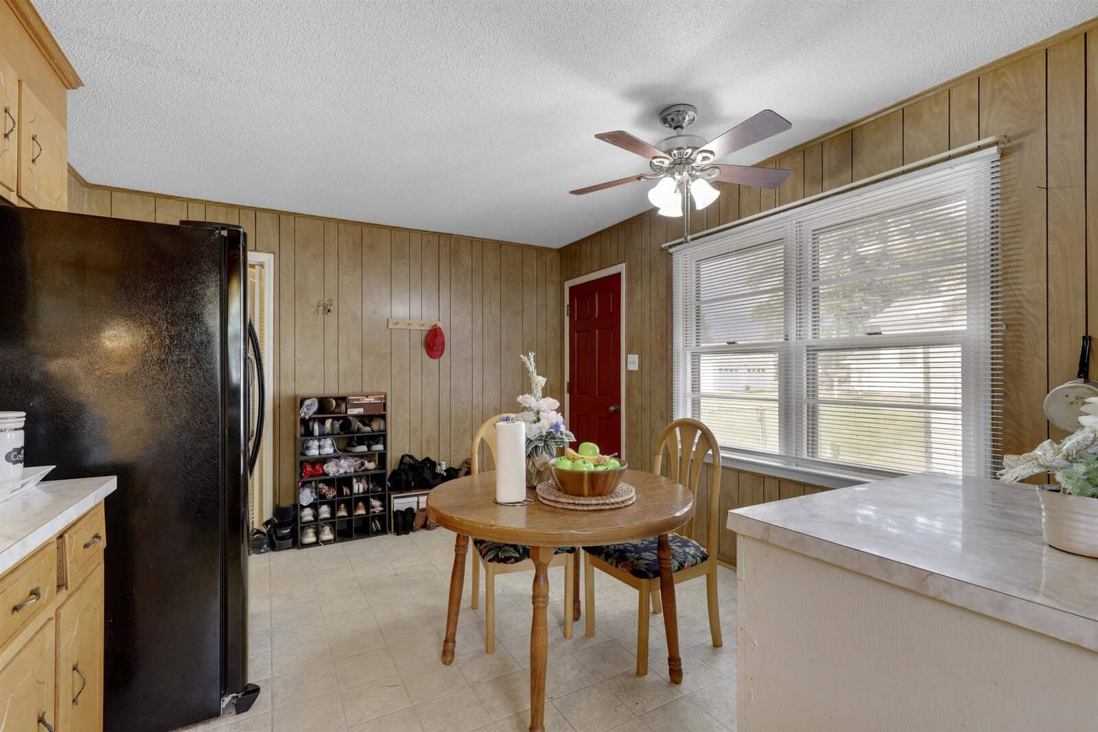 A modest kitchen and dining area with wooden paneling, featuring a refrigerator, shoe rack, a round wooden table with chairs, and a ceiling fan. natural light streams in through a window next to the red entry door.