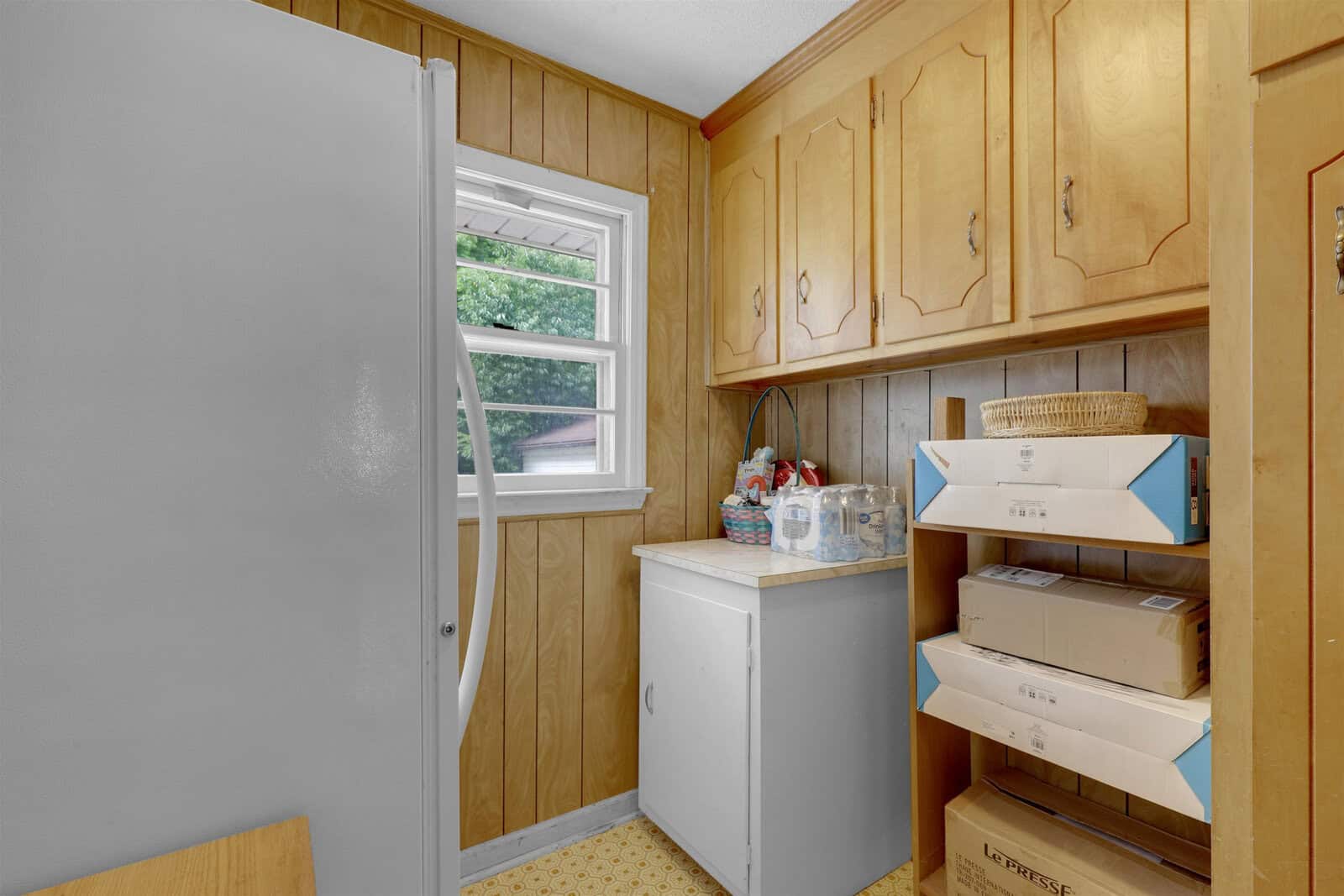 A modest laundry room featuring a top-loading washing machine, wooden cabinets, and a paneled window, with various laundry supplies and a wicker basket on the shelves.