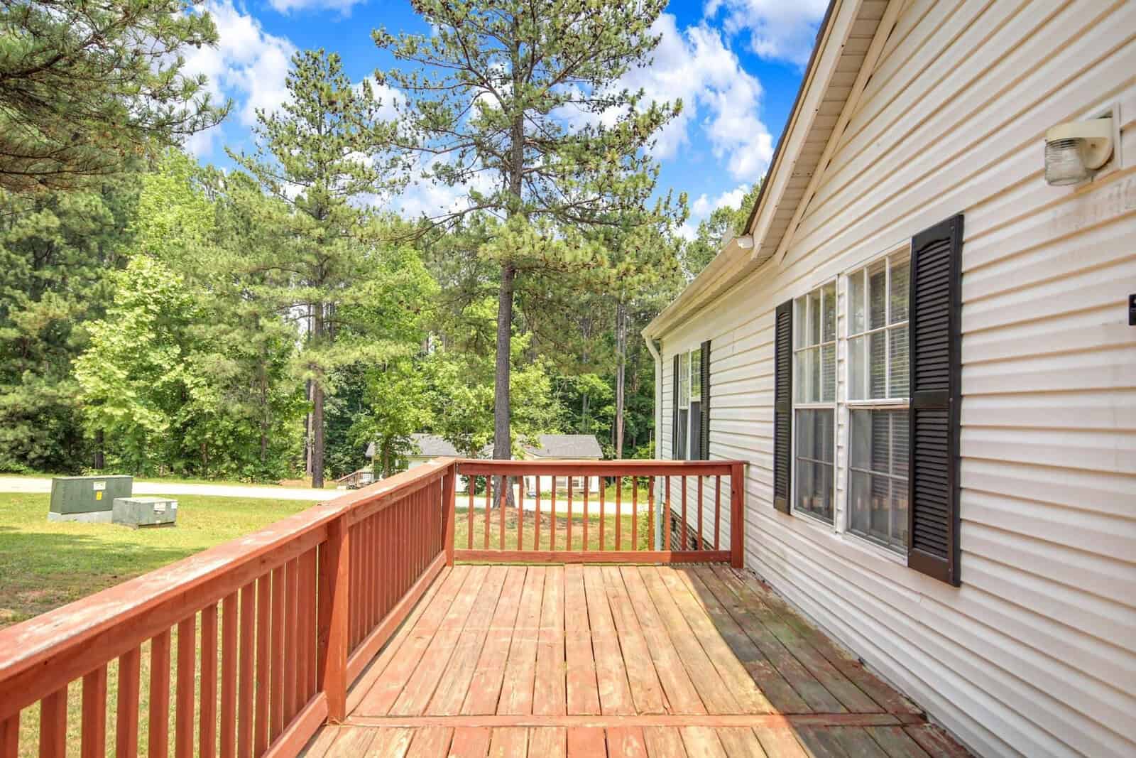 A wooden deck attached to a white siding house with windows, overlooking a yard with scattered trees and a clear sky above.