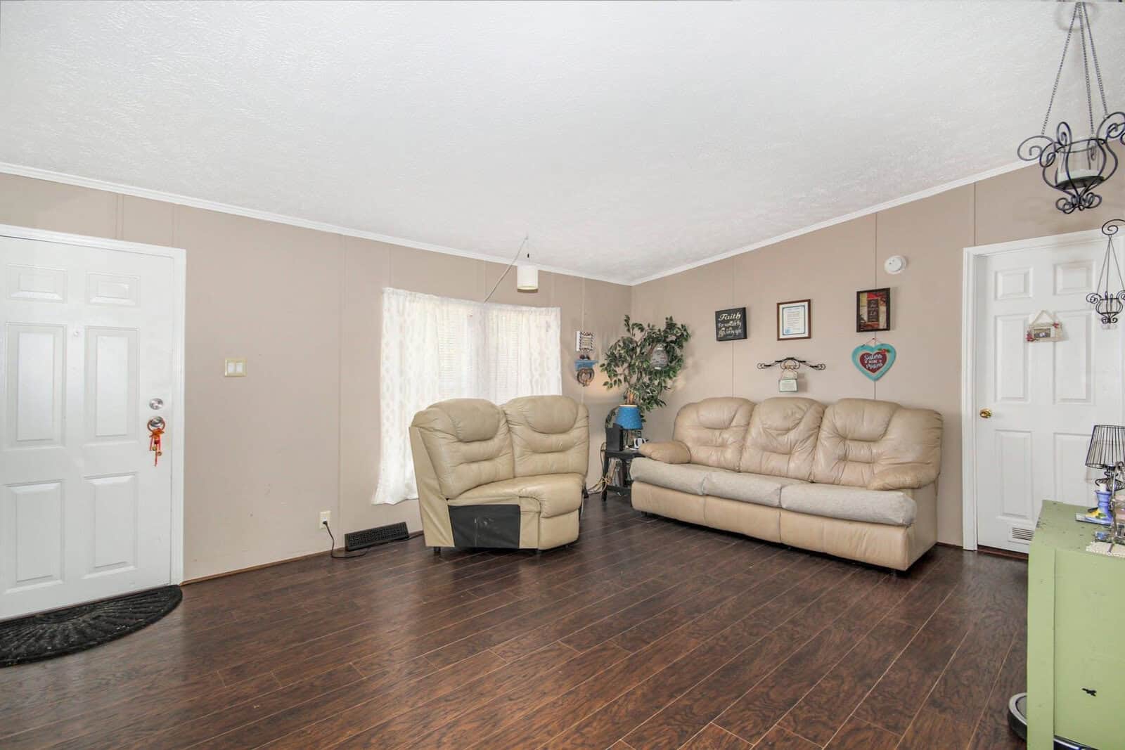 A living room featuring a cream-colored sofa set, wooden flooring, a white entrance door on the left, and a window draped with sheer curtains on the right. decorative elements include wall-mounted frames and a small indoor plant.