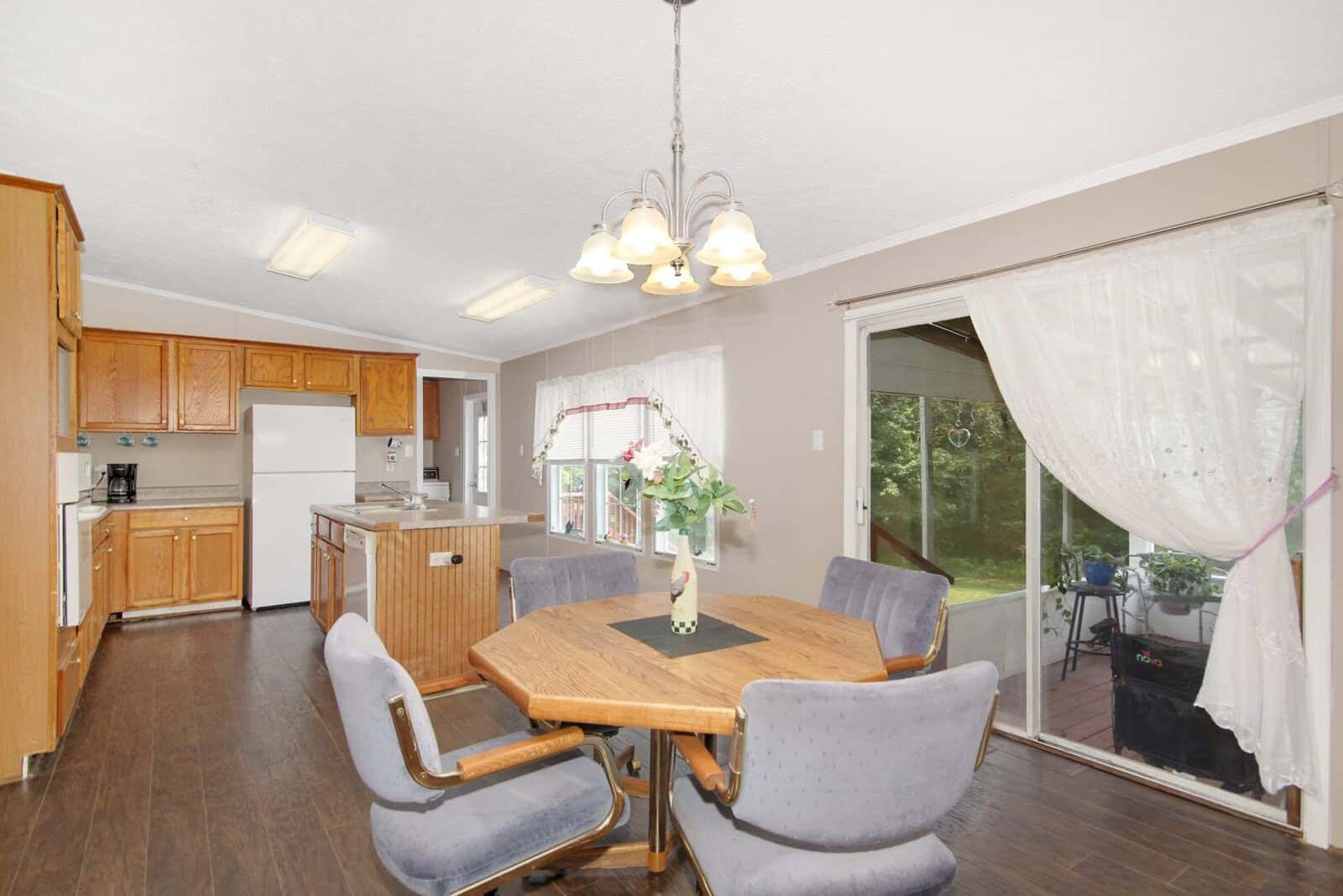 A well-lit dining area with a wooden hexagonal table, surrounded by six upholstered chairs, adjacent to a kitchen with wooden cabinets and white appliances, leading to a patio visible through a glass door.