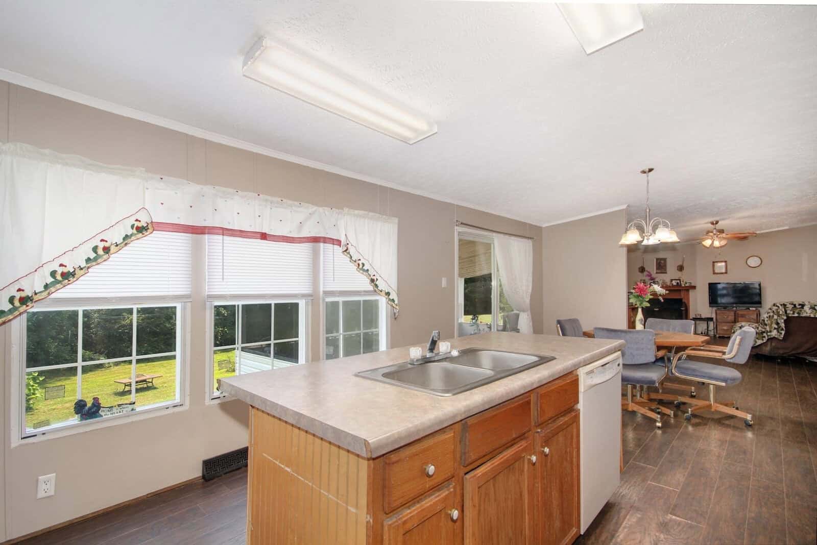Interior of a well-lit kitchen with wooden cabinets and a granite countertop, overlooking a dining area with a view of the backyard through the window.