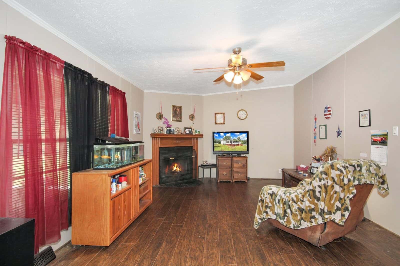 A neatly arranged living room with dark wood flooring, featuring a fireplace, a ceiling fan, a television set, and furnishings including a glass cabinet, a draped sofa, and red curtains.
