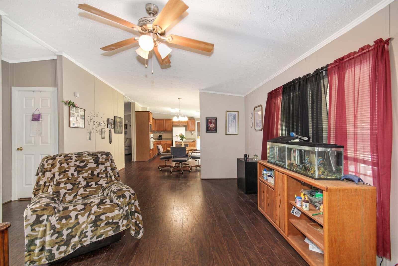 A well-lit living room with dark wood flooring featuring a camouflaged armchair, ceiling fan with lights, red curtains, an aquarium on a wooden stand, and an open plan view into the kitchen.
