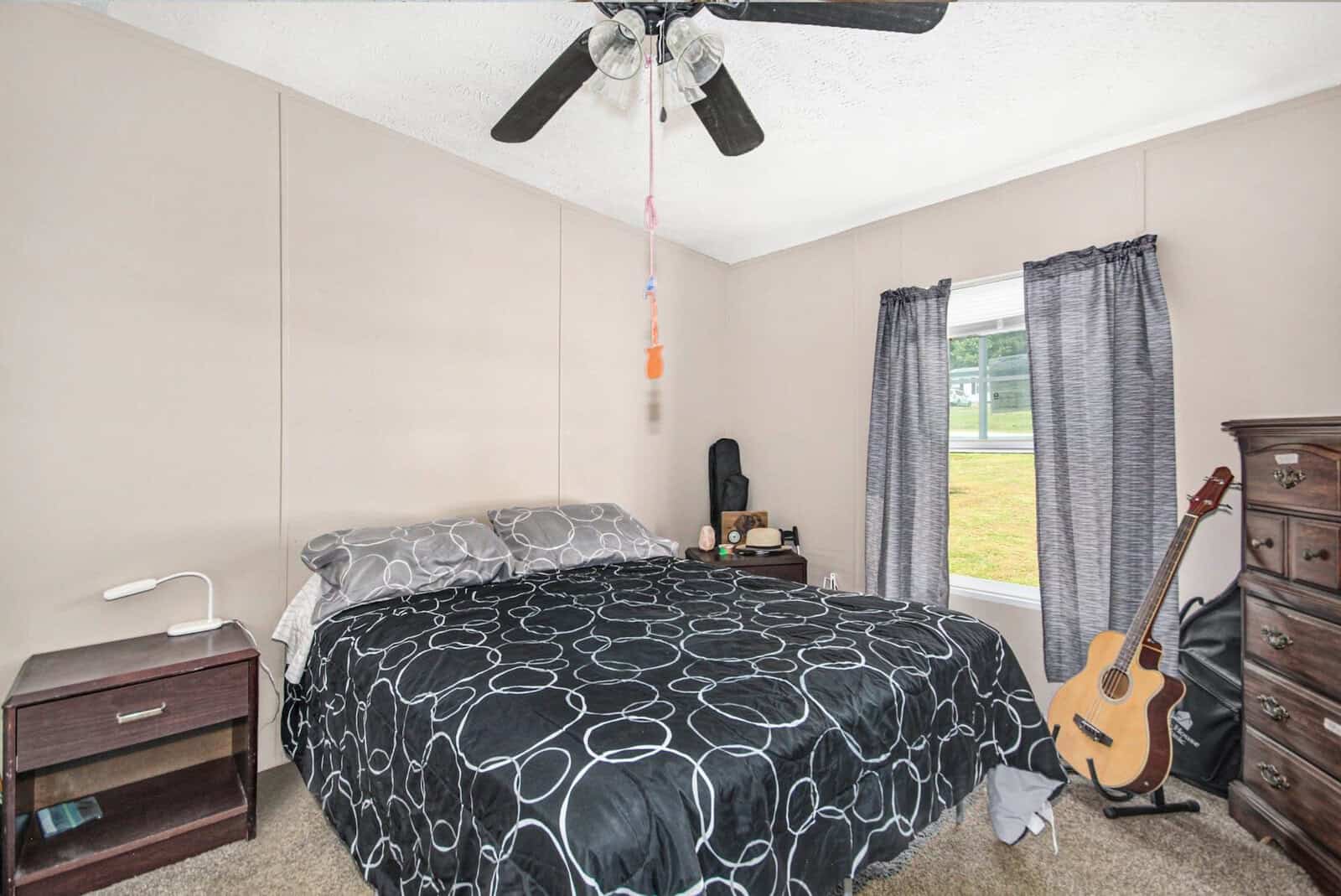 A modestly furnished bedroom with a full-sized bed covered in a black and white patterned comforter, a ceiling fan above, one acoustic guitar leaning against a wall, and a window with gray curtains. to the side, there are a nightstand and a small dresser with a few items on top.