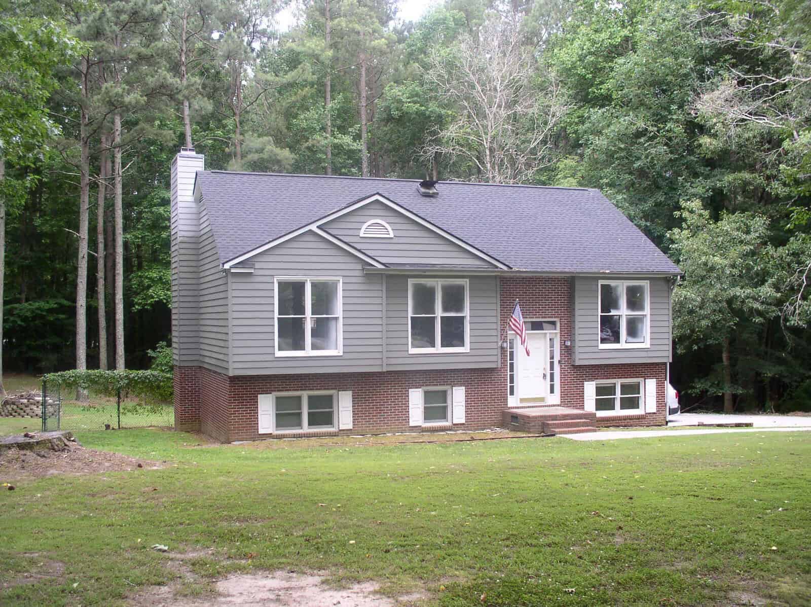 A two-story house with gray siding and red brick foundation situated in a clearing surrounded by trees at 57 Lakewood Court, Henderson NC. There is an American flag displayed on a pole attached to the