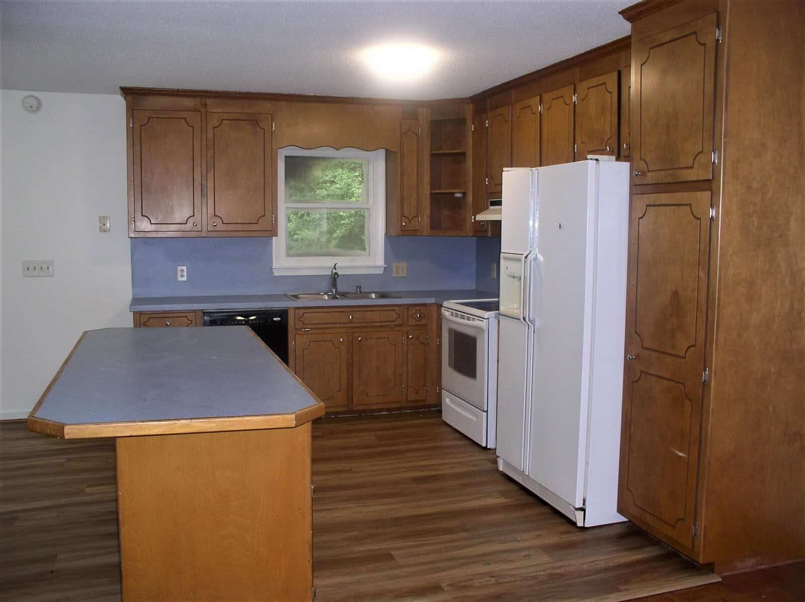A modest kitchen with wooden cabinets, blue walls, a central island, and white appliances including a refrigerator and a stove.