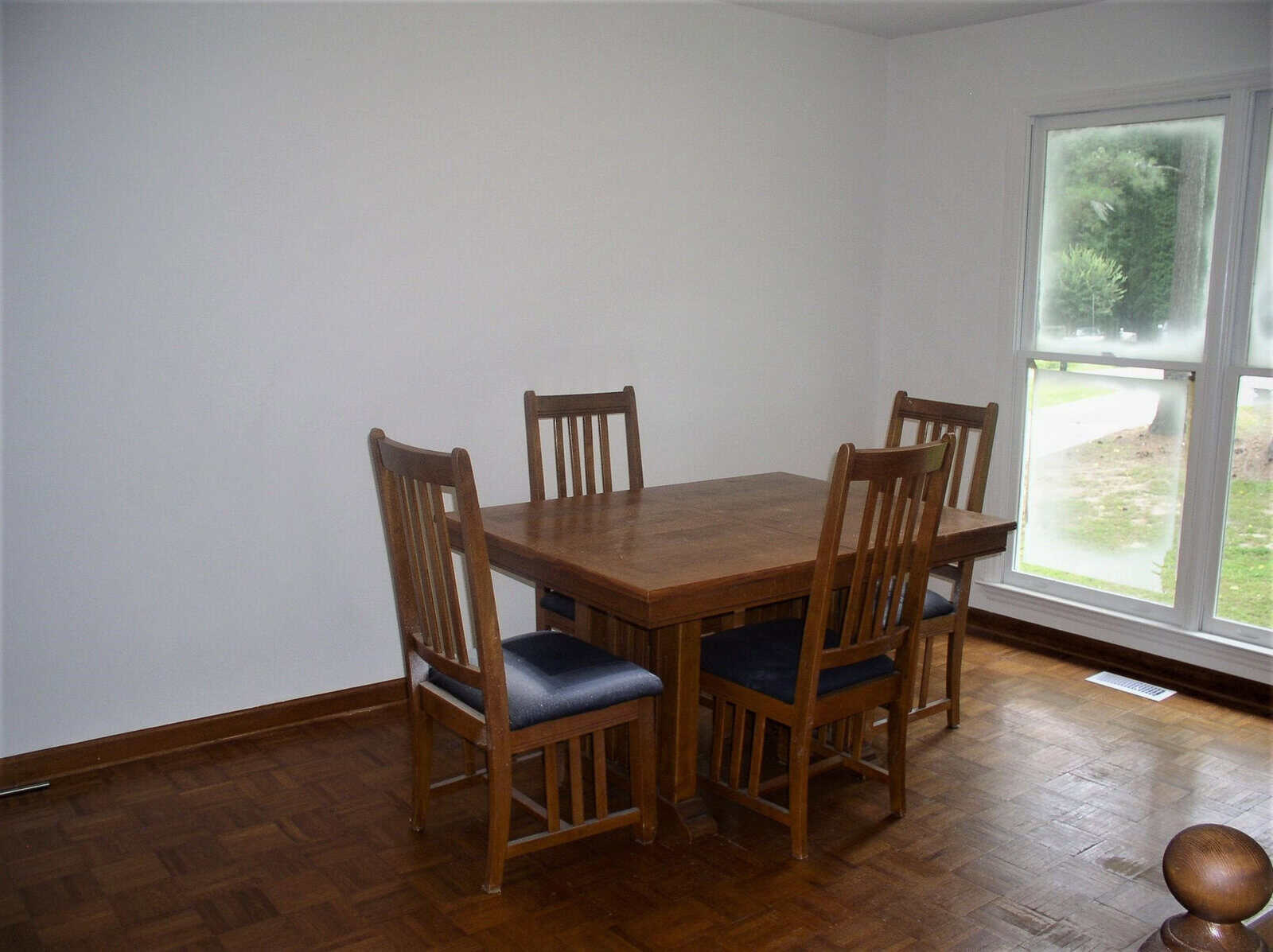 A wooden dining table with four chairs set in a room with hardwood floors, near a window overlooking a lawn.
