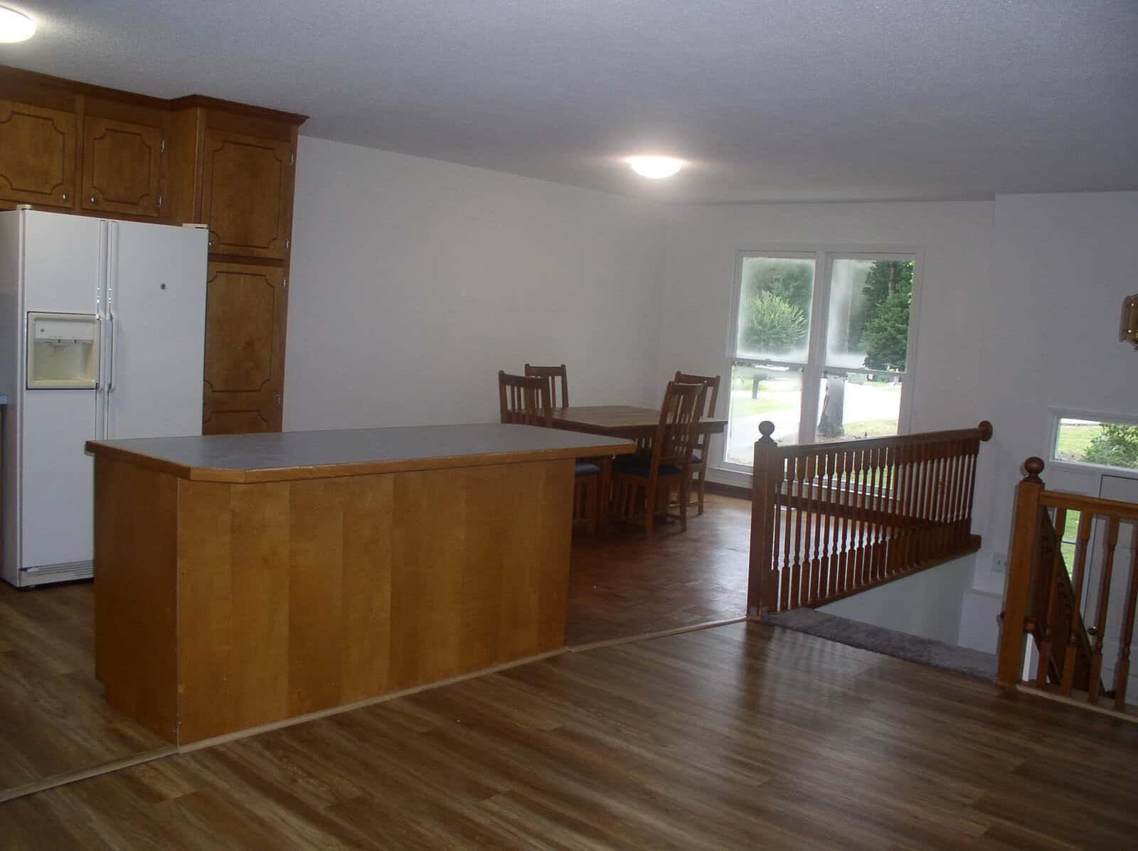 A spacious, unfurnished kitchen with wooden cabinets and an island, adjacent to a dining area with a simple table and chairs, overlooking a yard through a large window.
