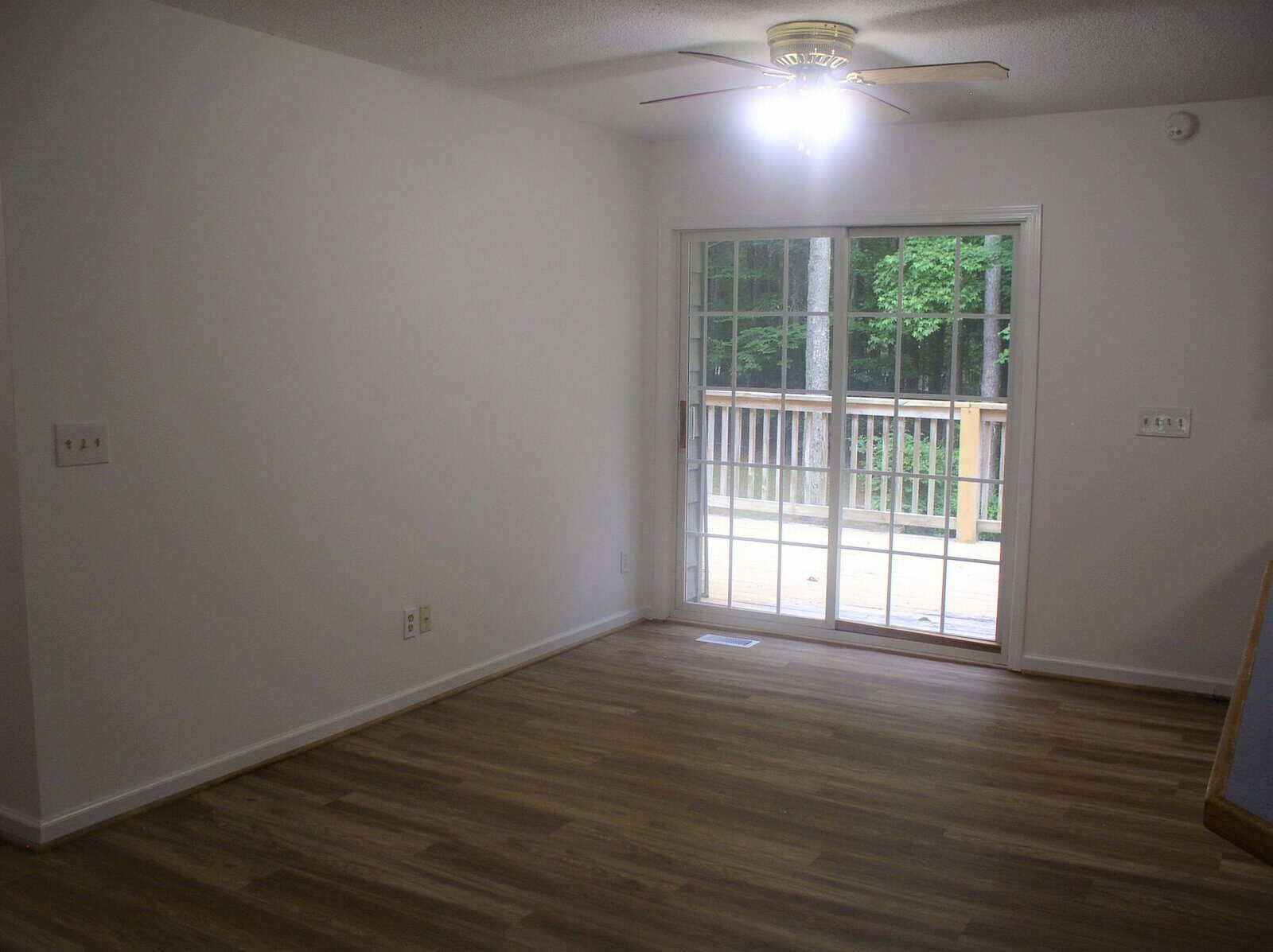 An empty room with laminate flooring, white walls, and a sliding glass door leading to an outdoor deck. there is a ceiling fan with lights in the center of the room.