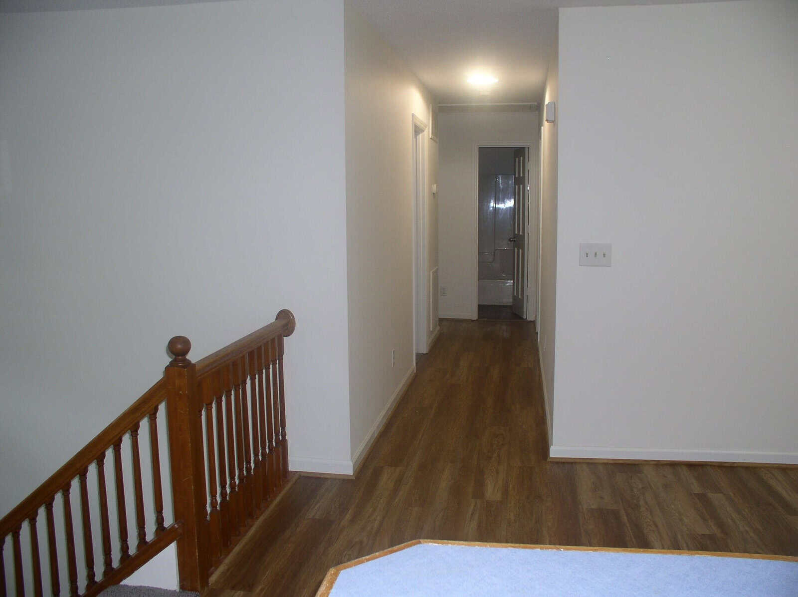 A sparsely furnished hallway with wooden floors, white walls, and a wooden railing to the left.