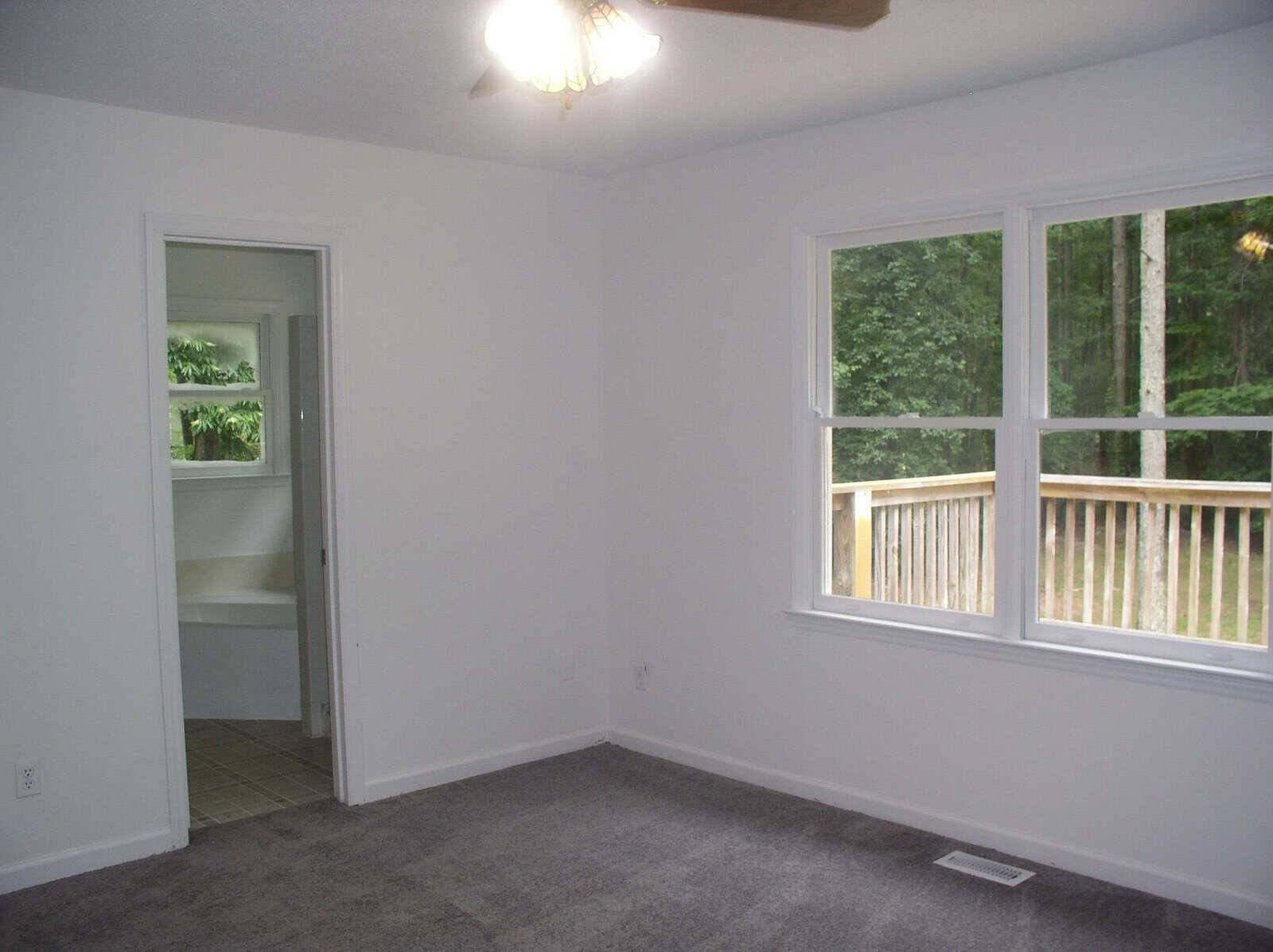 An empty room with gray carpeting, white walls, a ceiling fan, and two windows overlooking a wooden deck and trees. an open door reveals a glimpse of a tiled area.