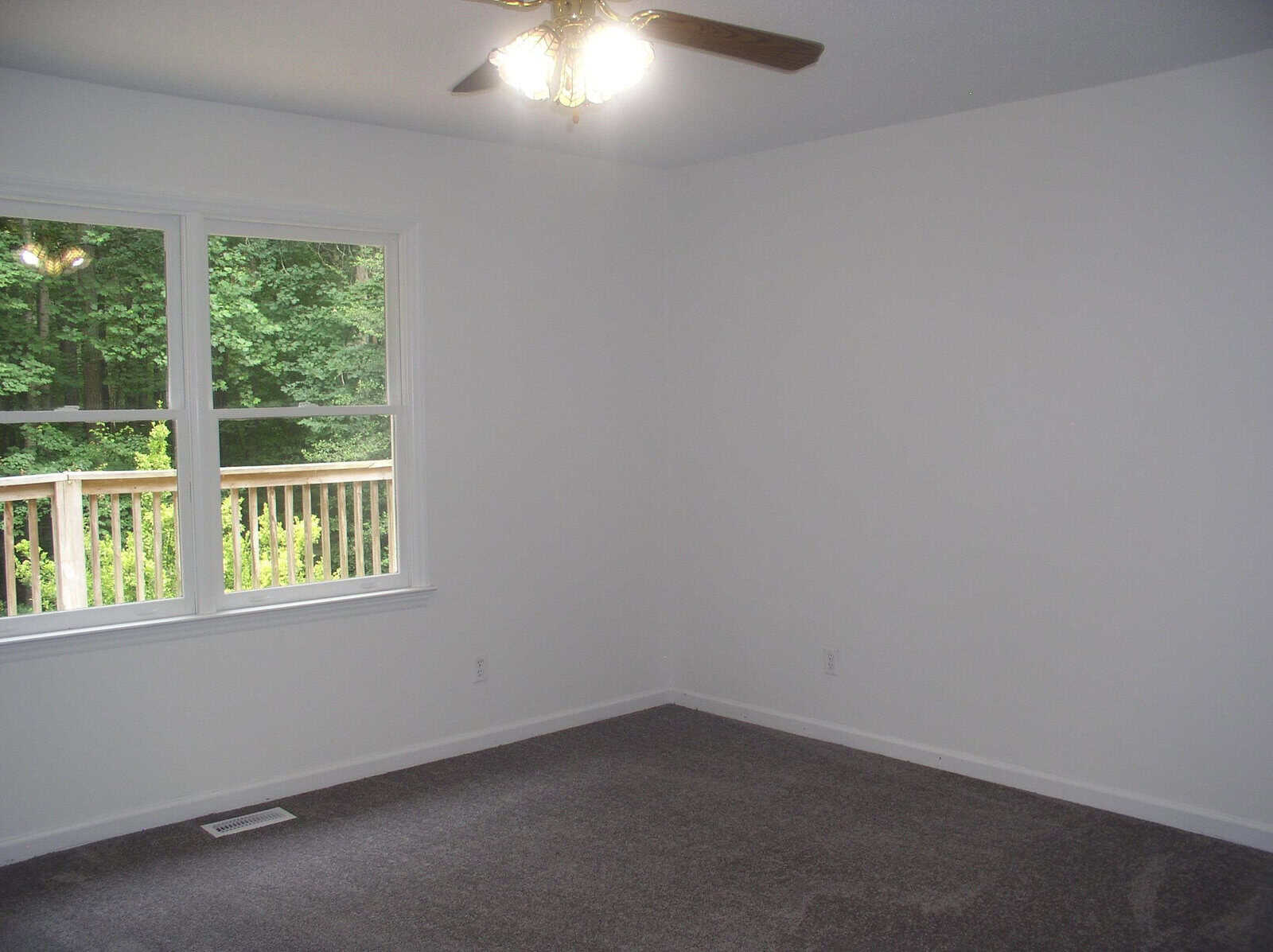 An empty room with white walls, a gray carpet, a ceiling fan with lights, and a window overlooking a wooded area with a deck.