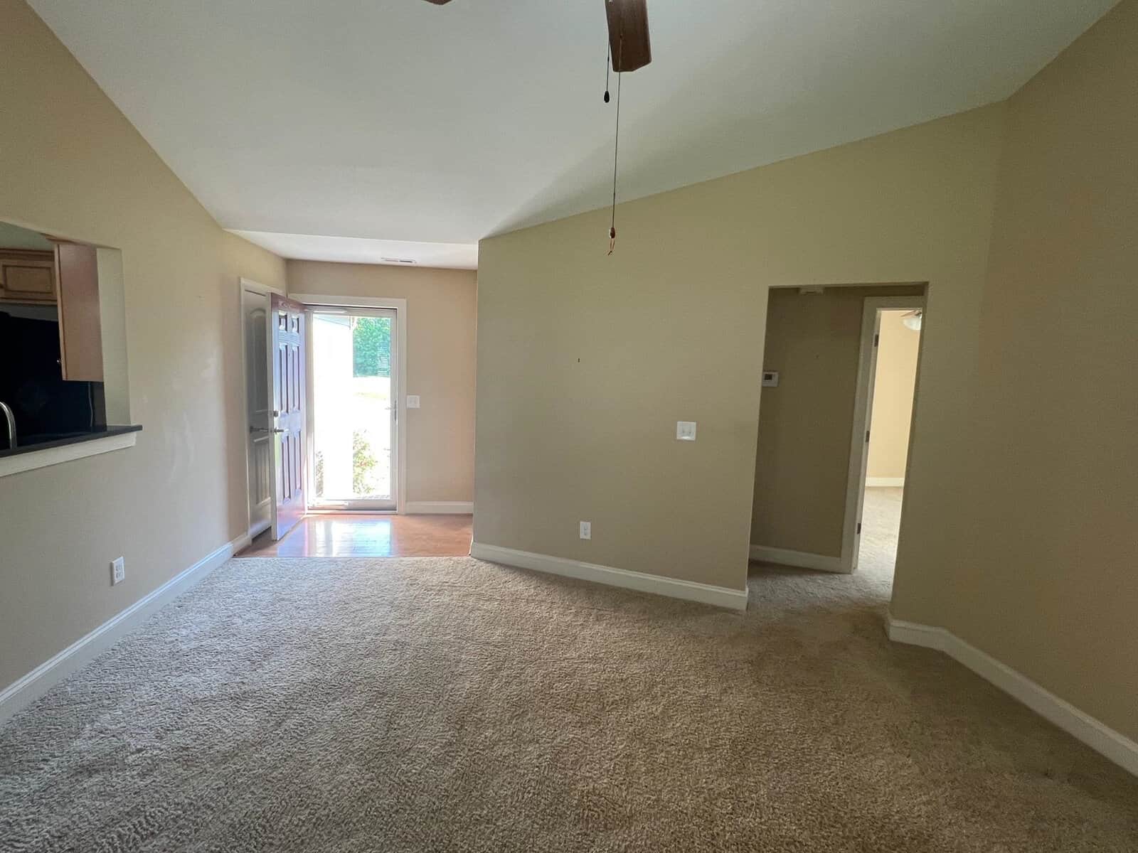 Empty living room with beige walls, carpeted flooring, and a sliding door leading outside.