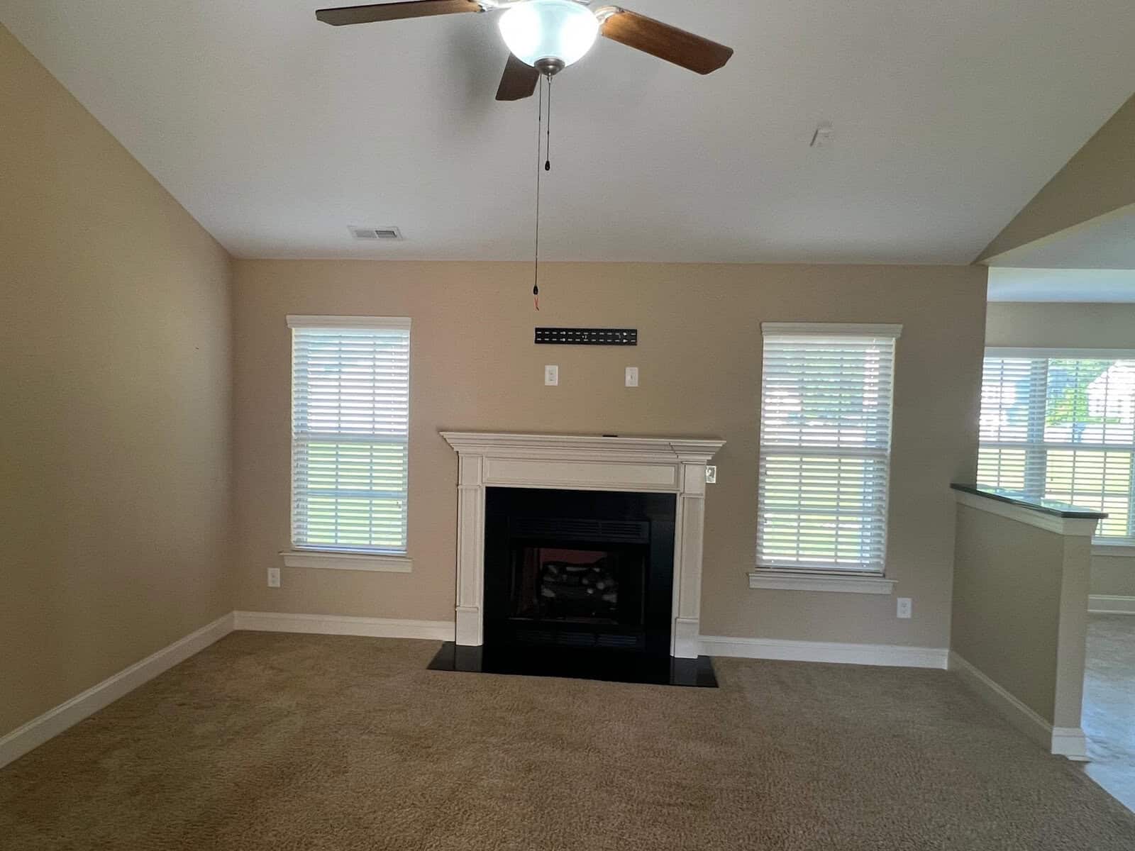 A spacious room featuring a fireplace with a white mantel, beige walls, two windows with white blinds, and a light brown carpet. a ceiling fan with wooden blades is installed on the high ceiling.