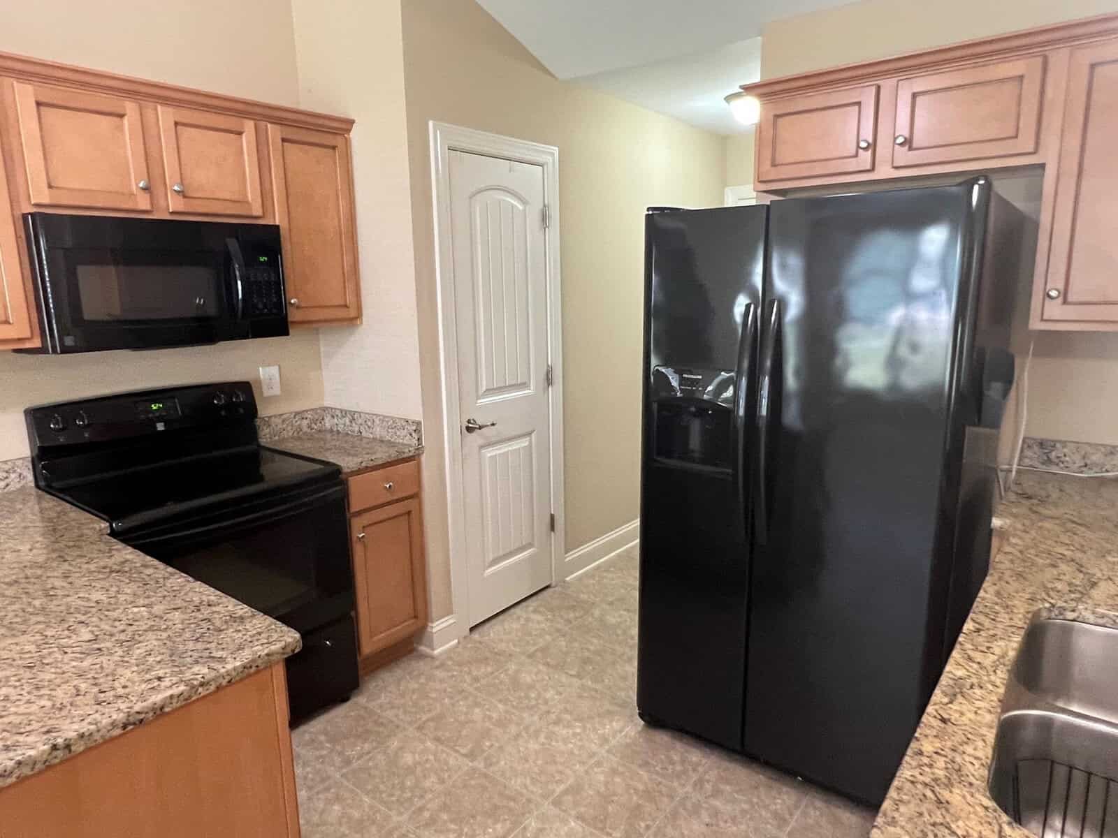 A residential kitchen with wooden cabinets, black appliances including a refrigerator and microwave, a stove, and beige tiled flooring. a door is visible in the background.