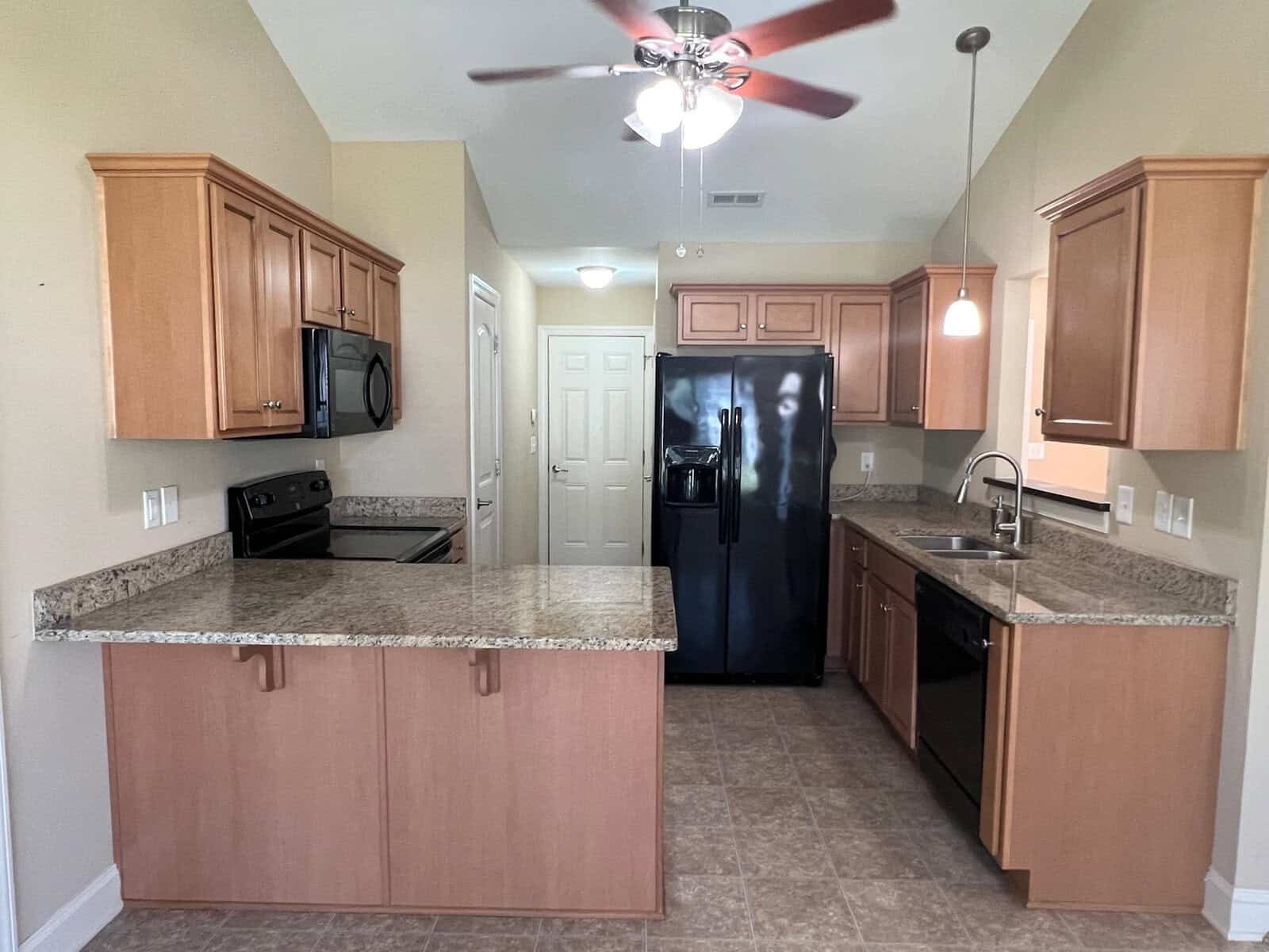 A modern kitchen interior with wooden cabinets, granite countertops, a black refrigerator, stove, and a ceiling fan.