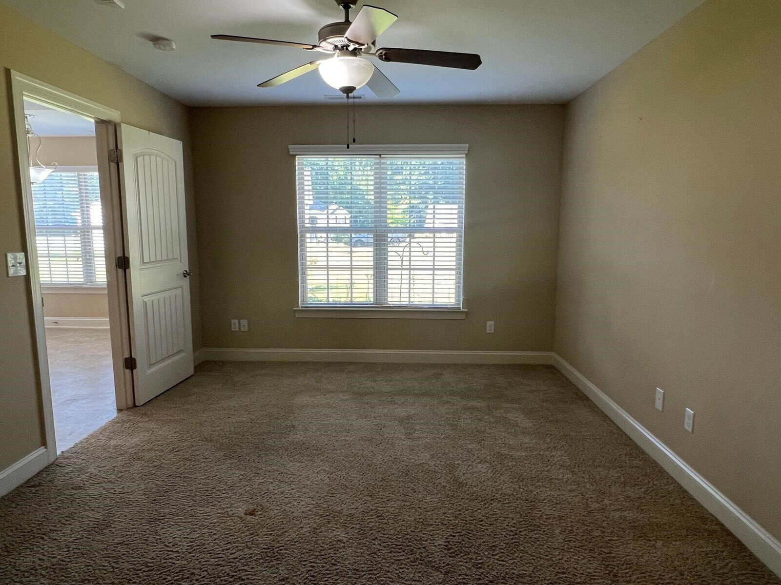 An empty room with beige carpeting, a single window with white blinds, and a ceiling fan with lights.
