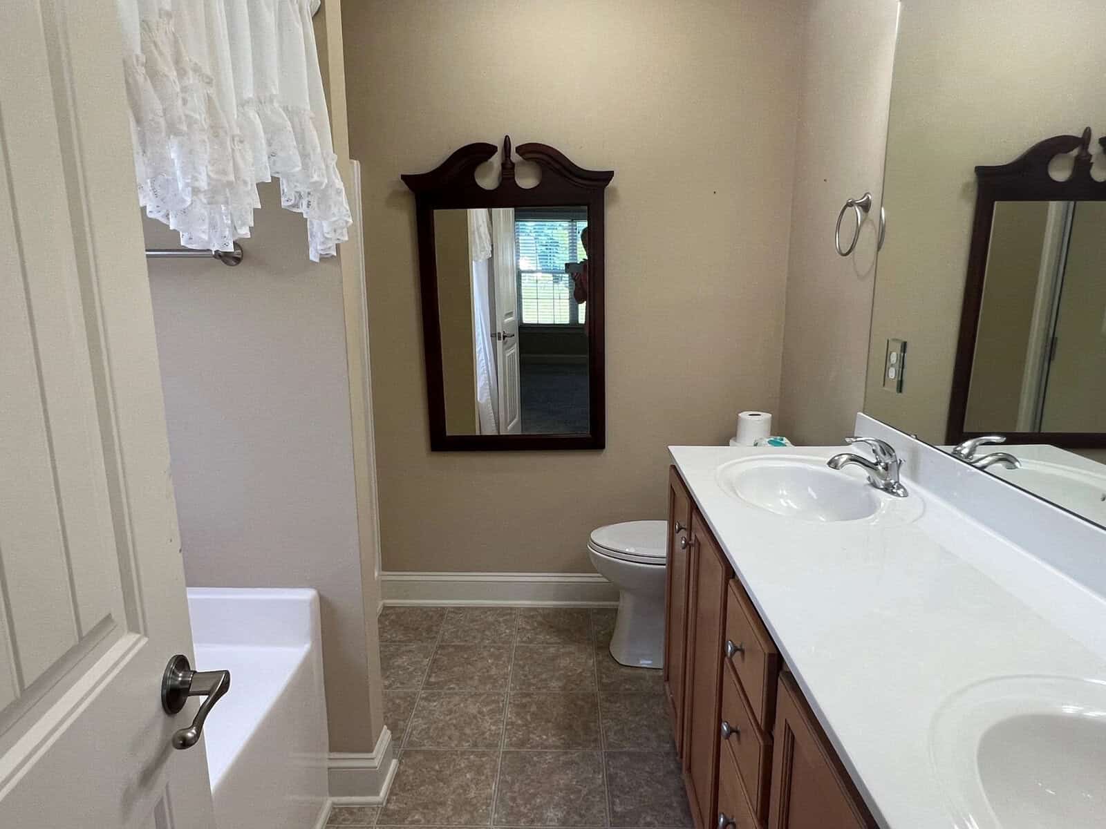 A residential bathroom featuring a dual sink vanity, two mirrors, a toilet, and a bathtub, with a window providing natural light.