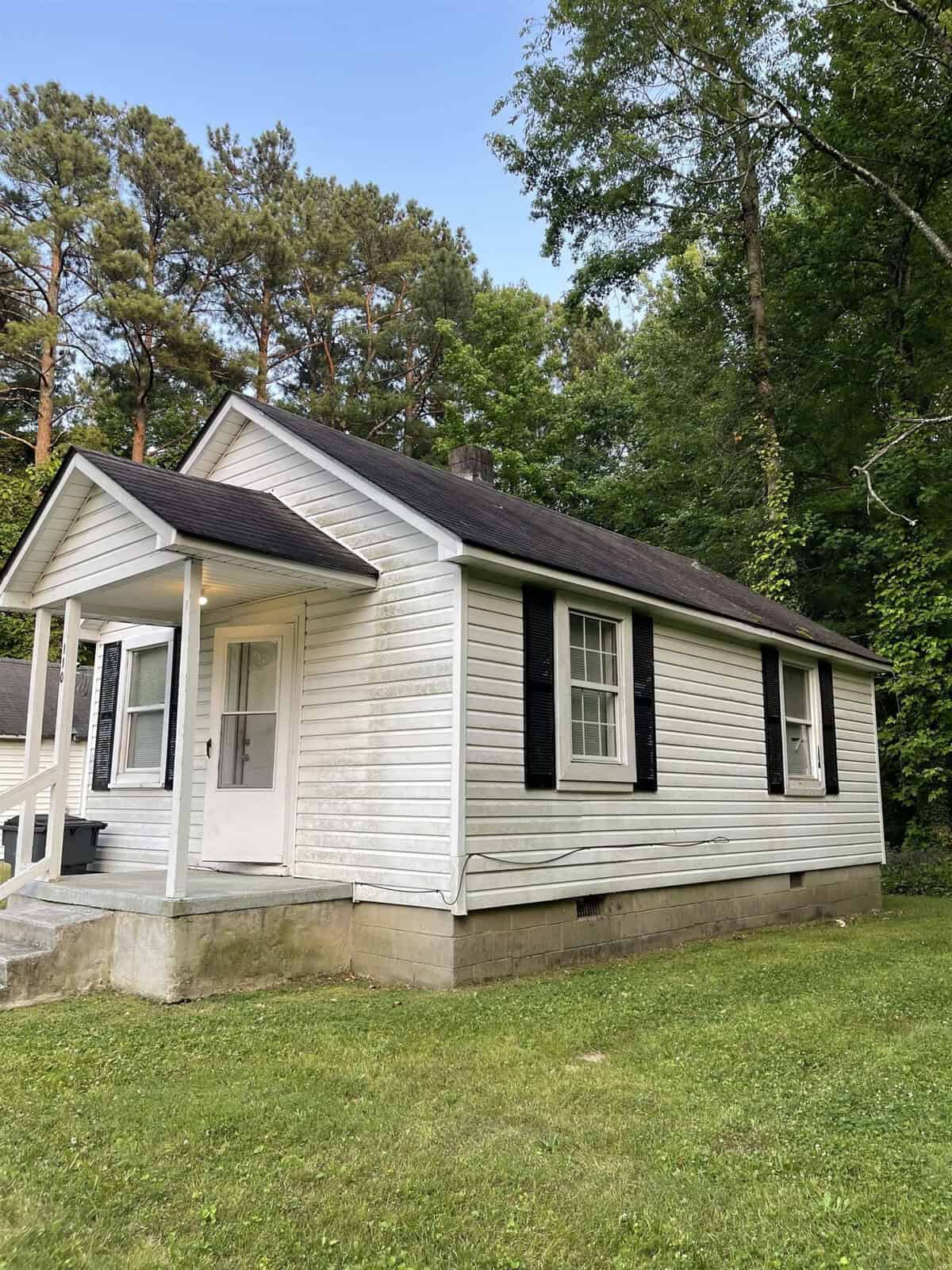 A small, white single-story house with a gabled roof and black shutters on the windows, located in a wooded area at 110 Cleveland Avenue, Henderson NC.