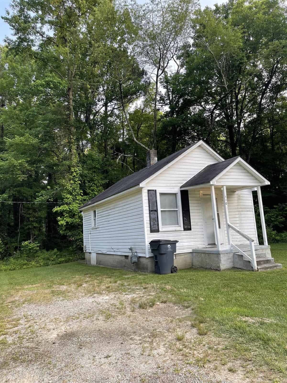 A small white single-story house with a gabled roof and an attached front porch is situated next to a wooded area, with a gravel space in the foreground and a black trash bin by the side.