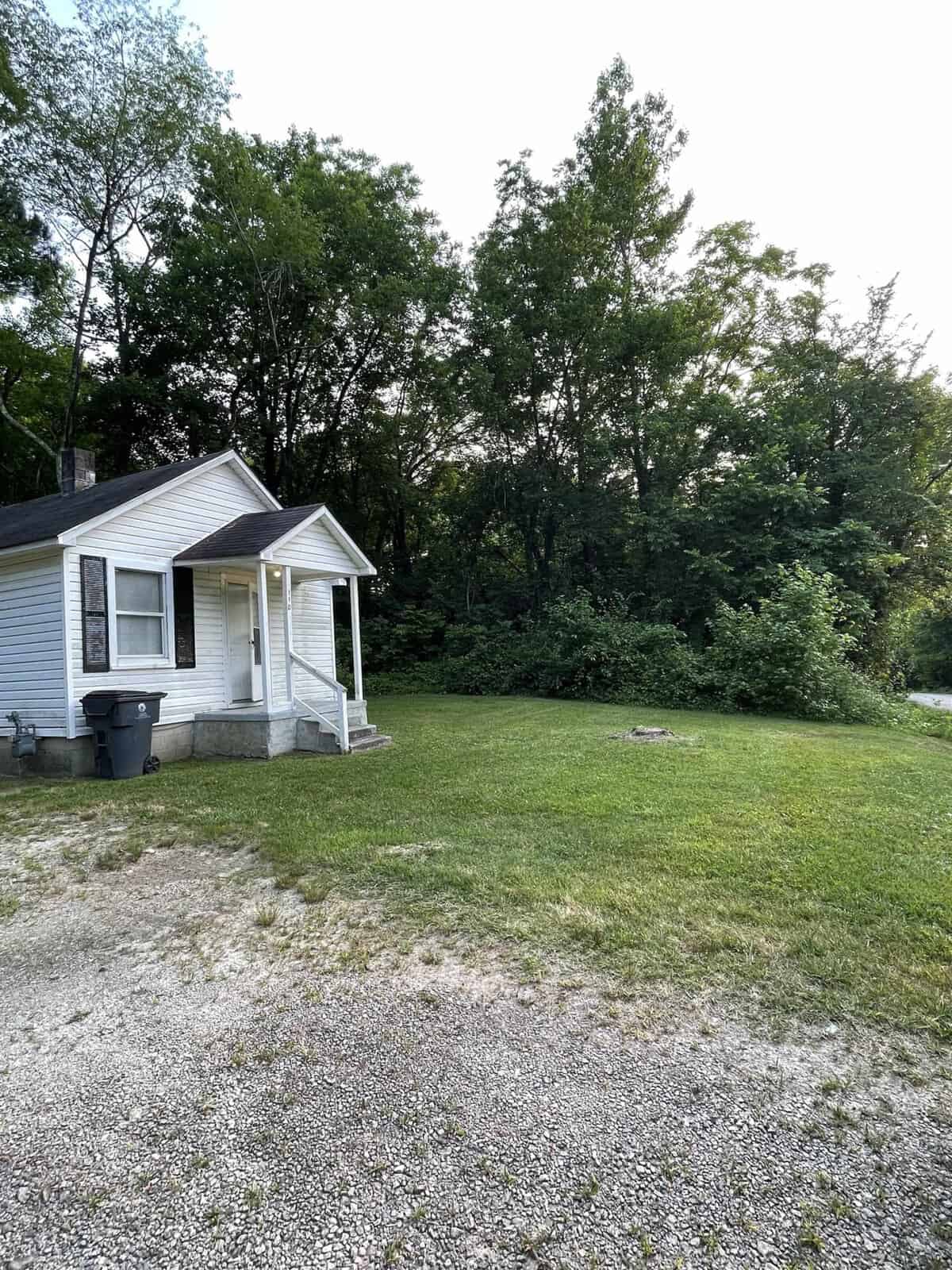 A small white house with a front porch situated next to a wooded area with a gravel driveway extending in the foreground.