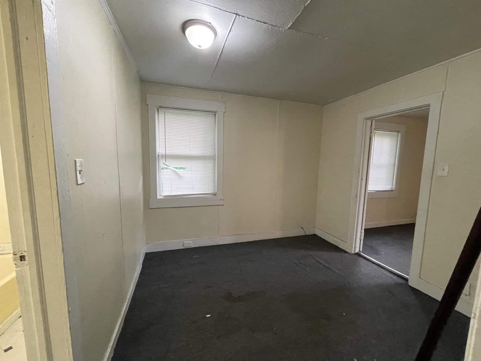 An empty room with white walls, a gray carpet, two windows with closed blinds, and one visible doorway.
