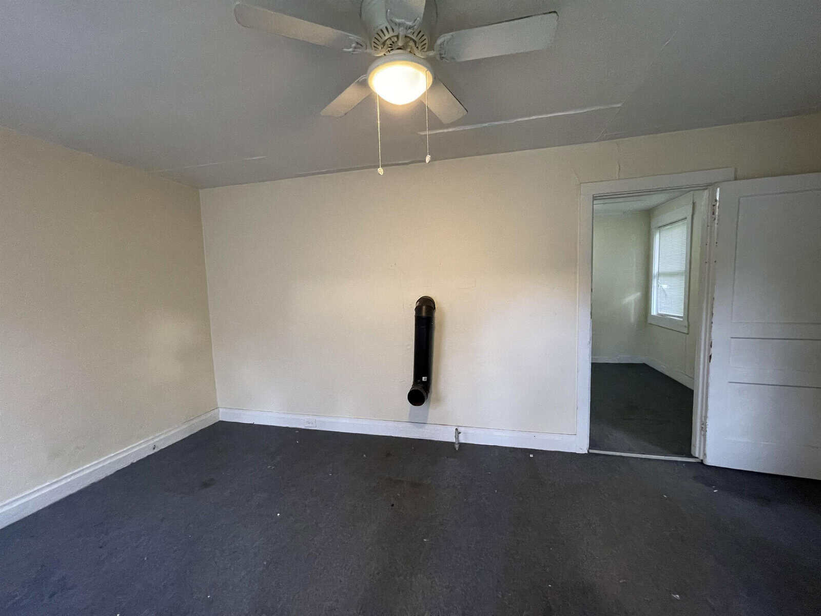 An empty room with white walls and a dark-colored carpet, featuring a ceiling fan and a singular, wall-mounted, black soundbar speaker.