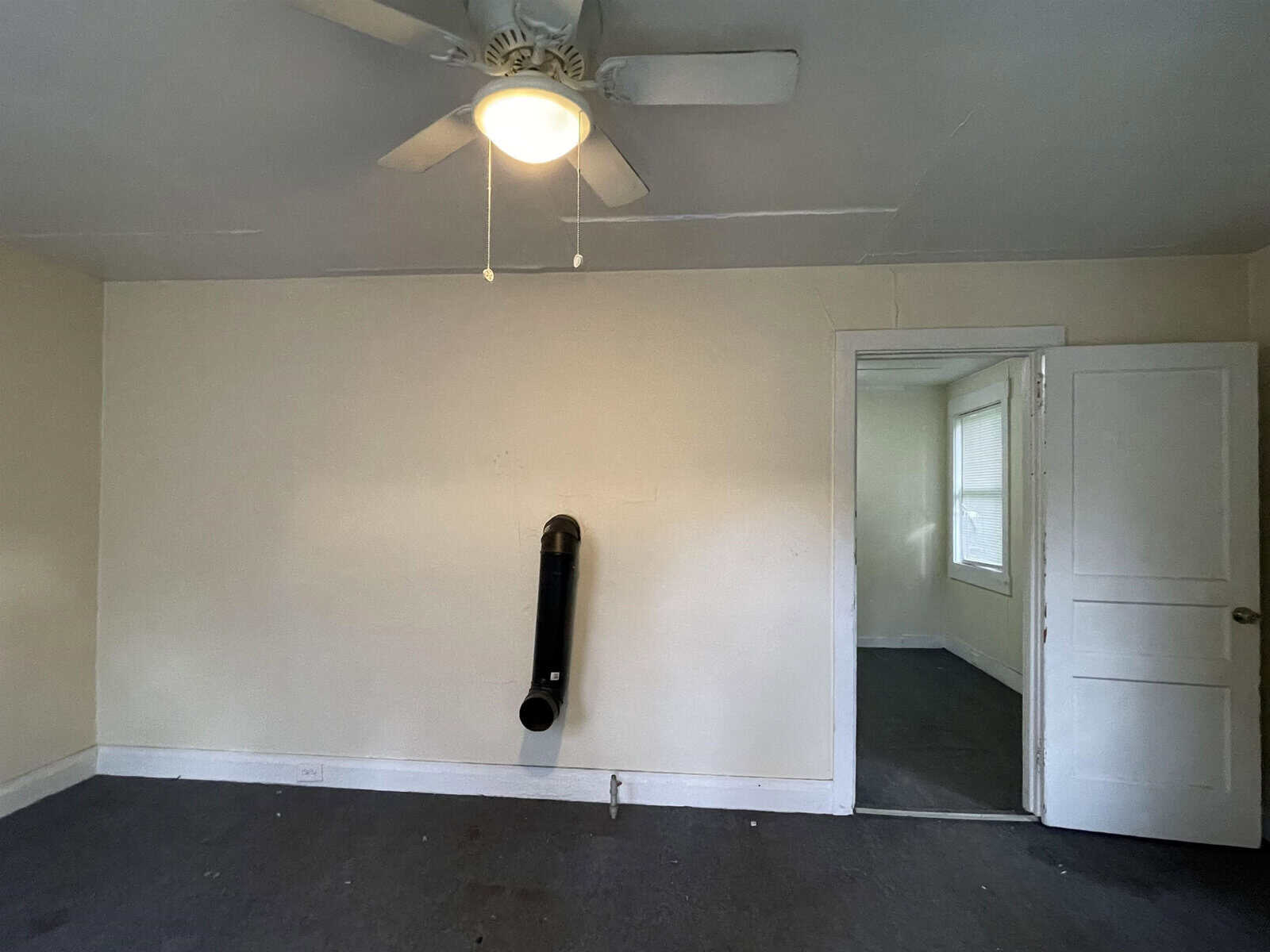 An empty room with white walls, a ceiling fan with lights, a black pull-up bar mounted on the wall, and a closed door on the right side.