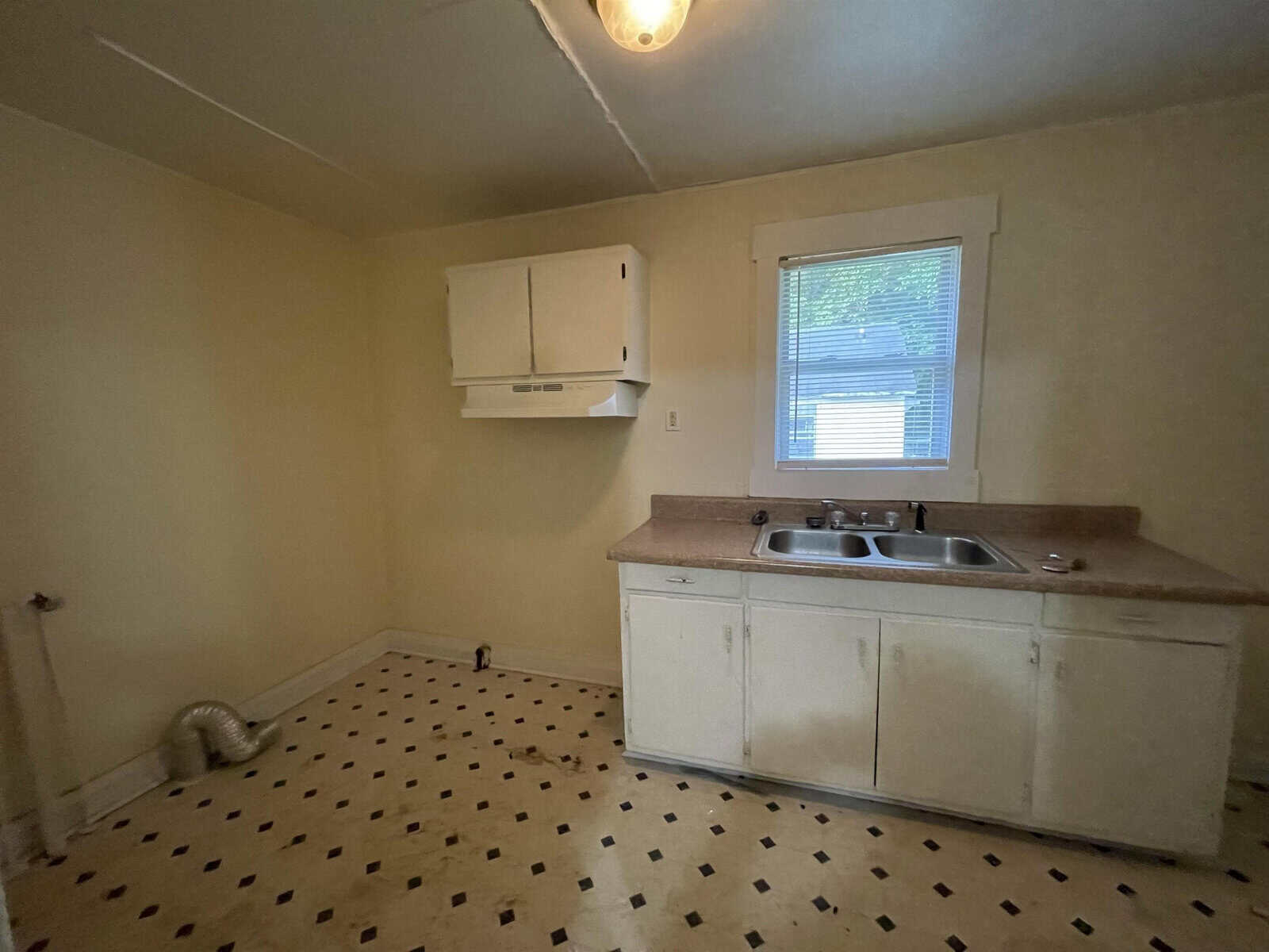 An unfurnished room with an old kitchenette comprising a sink and under-sink cabinets, alongside a wall-mounted cabinet, set against plain walls with a single window, and a patterned linoleum floor.