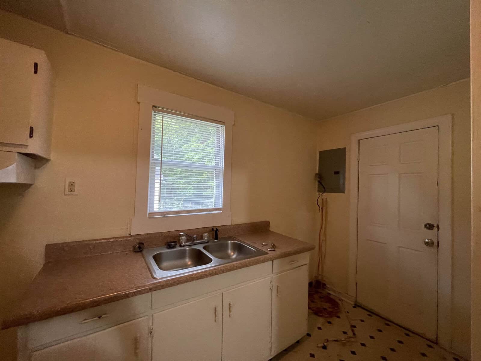 A sparse, outdated kitchen with a double sink, a small window, and a closed door. the walls and cabinets appear to be in need of maintenance or renovation.