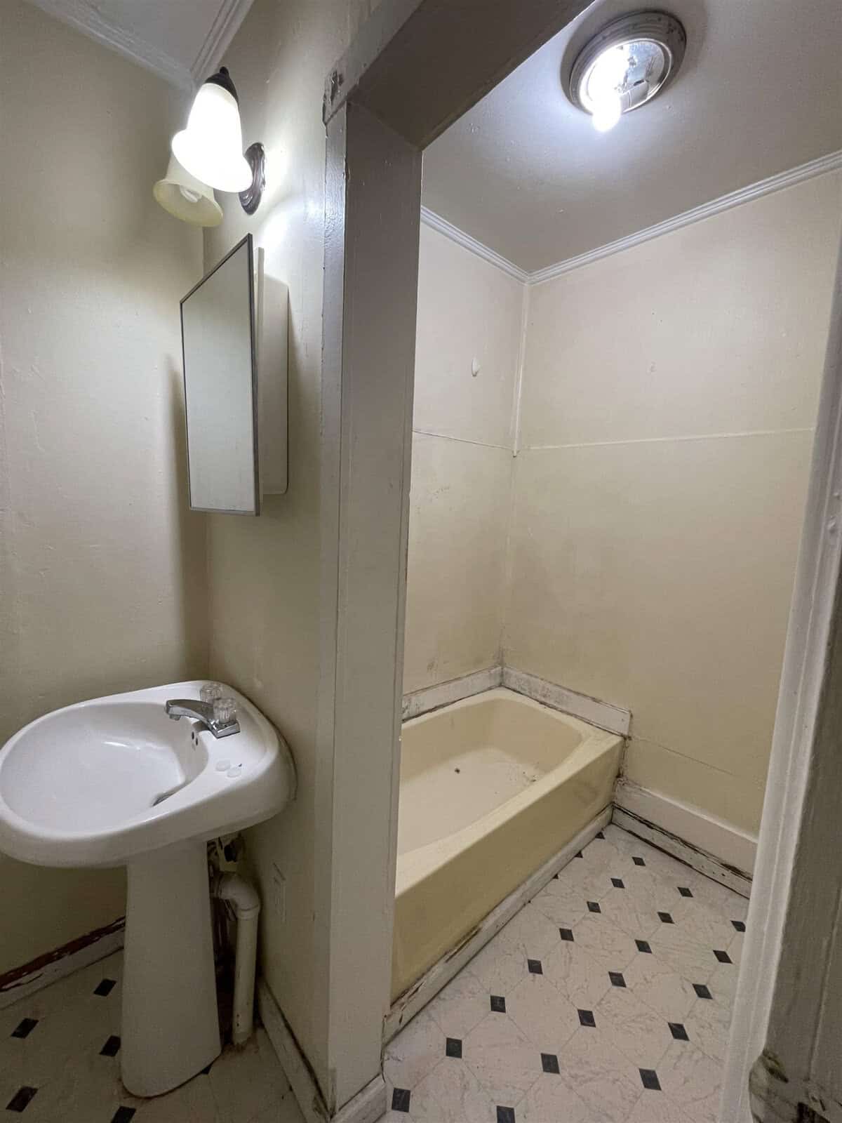 A small, cluttered bathroom with a pedestal sink, a corner bathtub, tiled flooring, and a mirror on the wall.