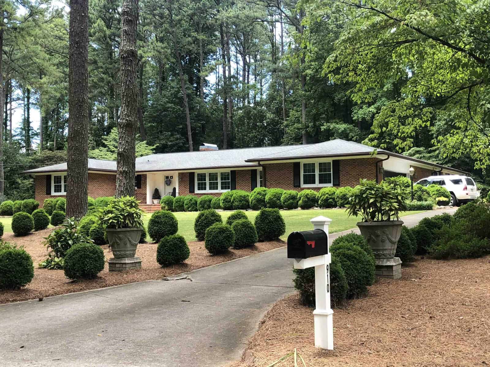 Single-story brick house with white trim, surrounded by manicured bushes and trees, featuring a curved driveway and a white mailbox in the foreground.