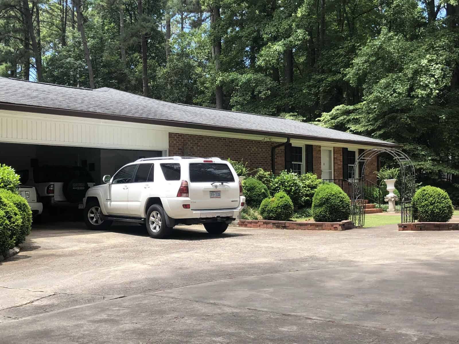 A white suv parked in the driveway of a suburban house with an open garage and surrounding greenery.