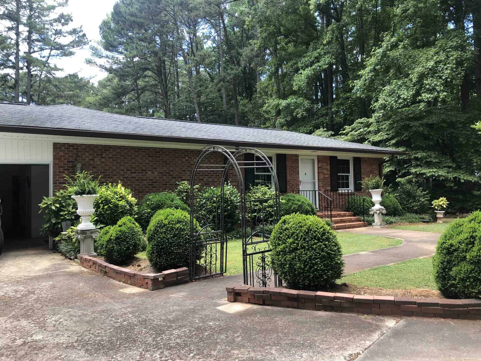 A single-story brick house with a white door, flanked by shaped bushes and decorative planters, featuring a garden arch near the walkway, set against a backdrop of tall trees.