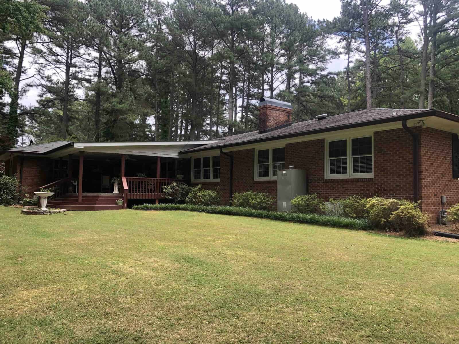 A single-story brick house with a front porch, surrounded by a well-manicured lawn and tall pine trees.