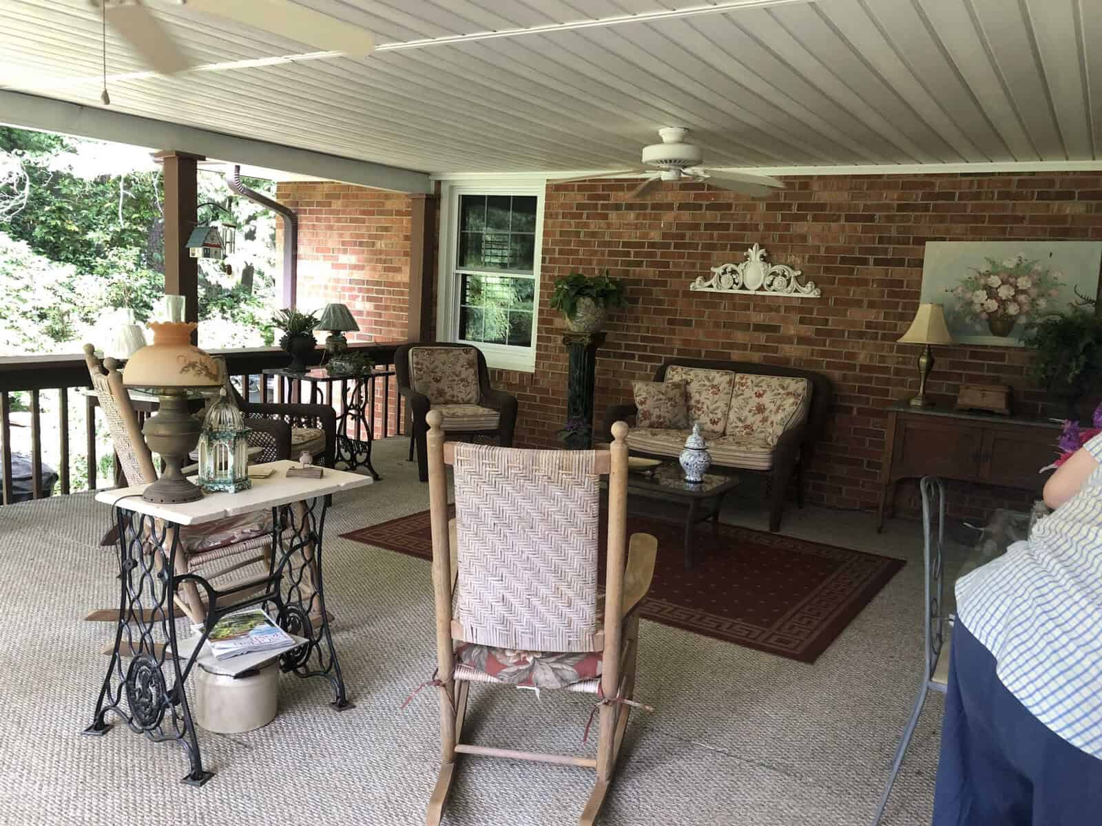A covered outdoor patio area with brick walls, furnished with a rocking chair, a small dining table, and a sitting area with upholstered chairs and decorative items.