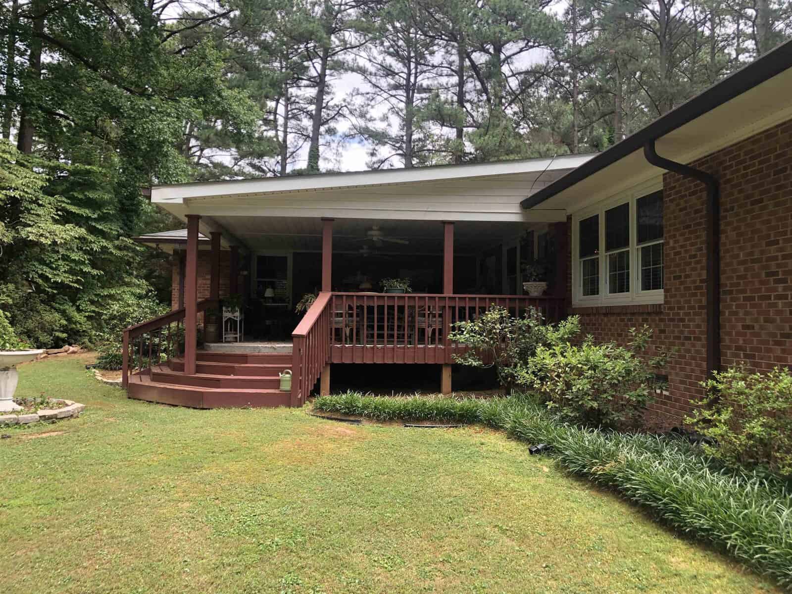 A one-story brick house with a covered wooden porch, surrounded by green lawn and tall pine trees.