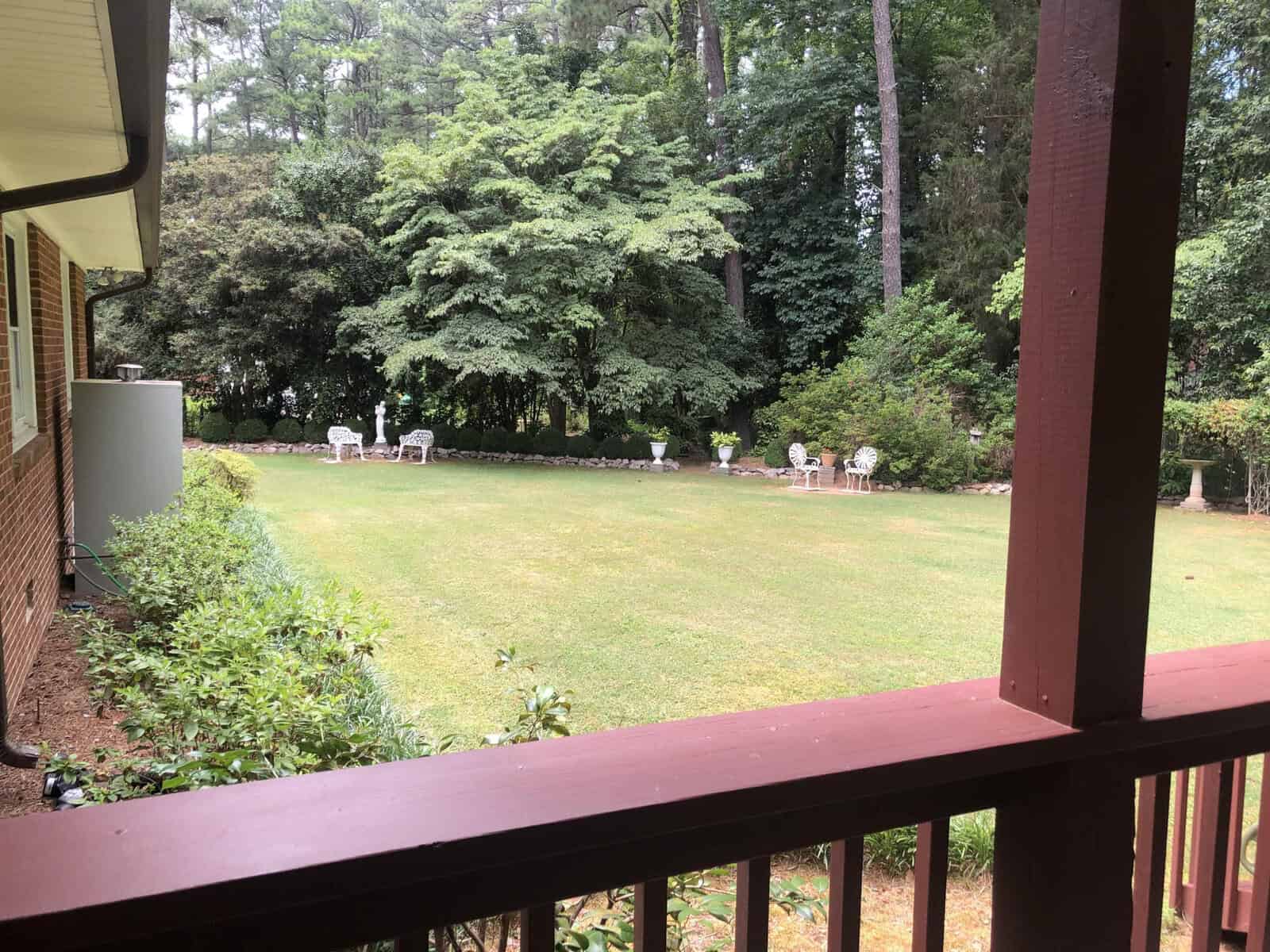 A view from a shaded porch overlooking a well-manicured lawn bordered by dense trees and shrubbery, with white outdoor chairs arranged in the distance.