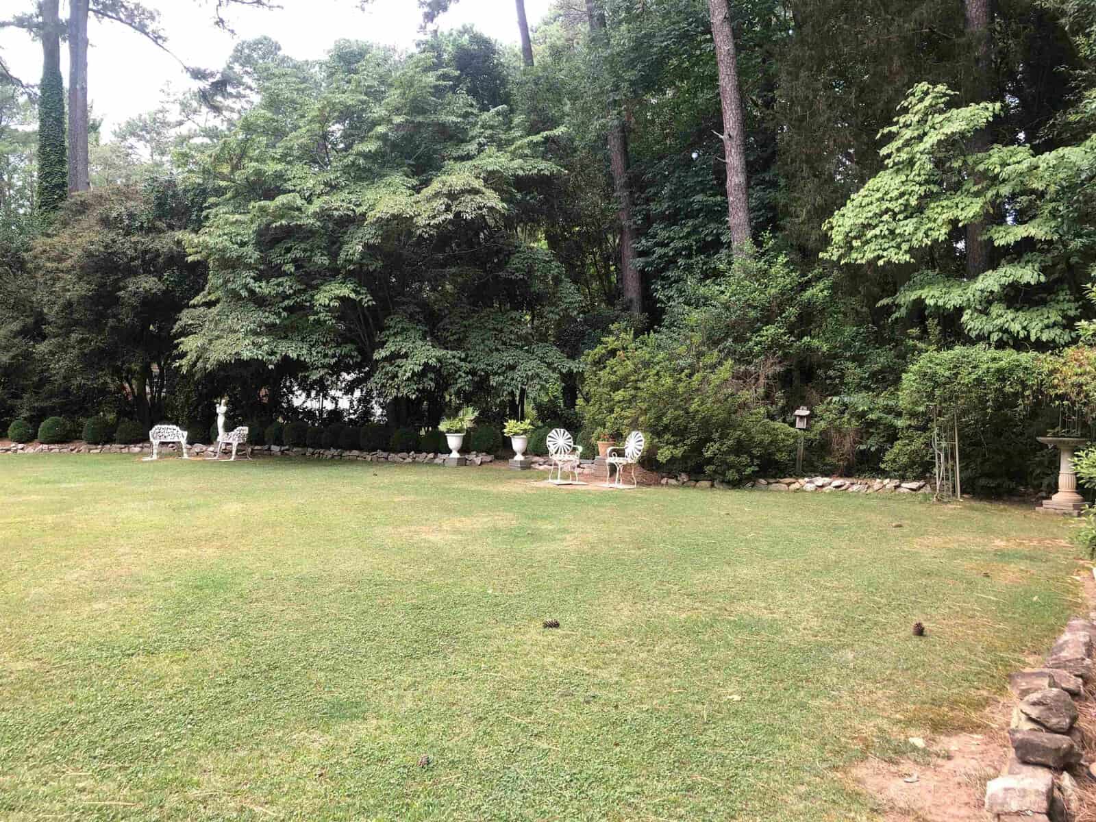 A grassy backyard with trees and a sitting area featuring white chairs and a table.