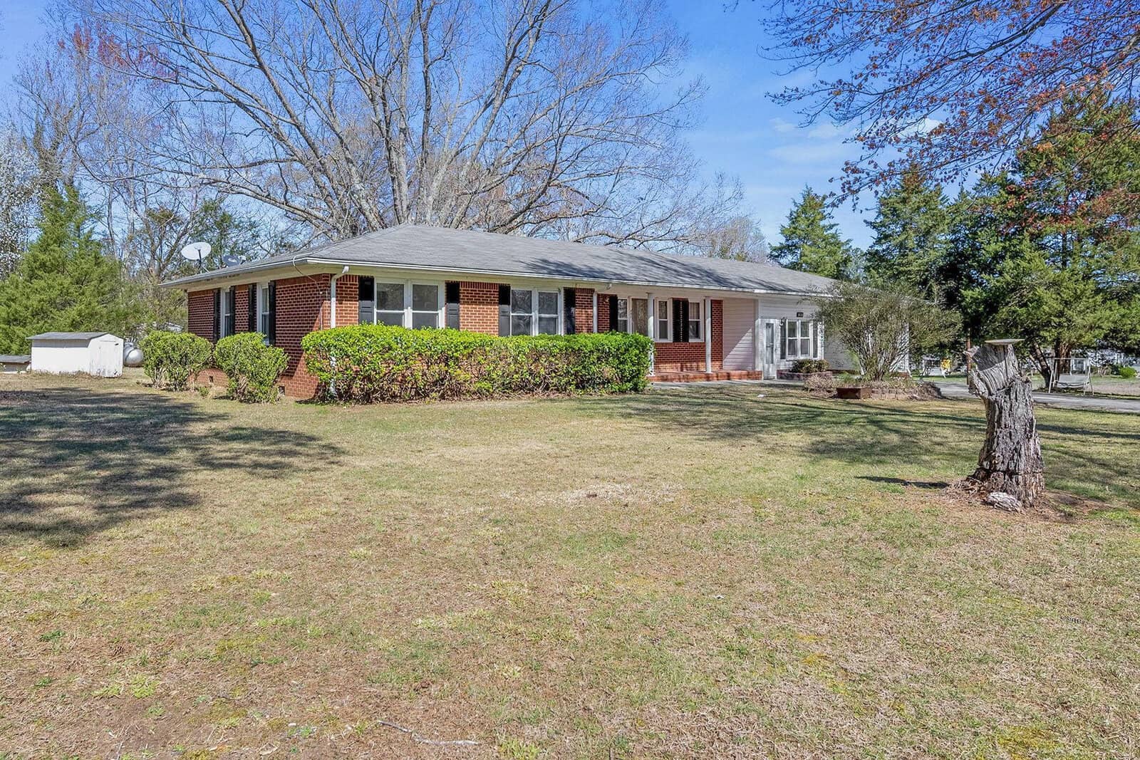 A single-story brick house with white trim, surrounded by a grassy lawn with a few trees and bushes, under a clear blue sky.